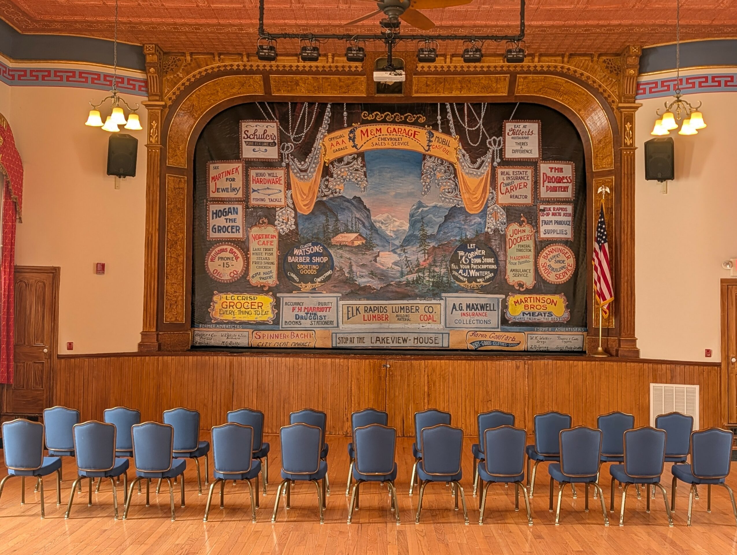 Empty chairs face a decorated stage with a mountain mural and vintage signs, in a theater or auditorium setting.