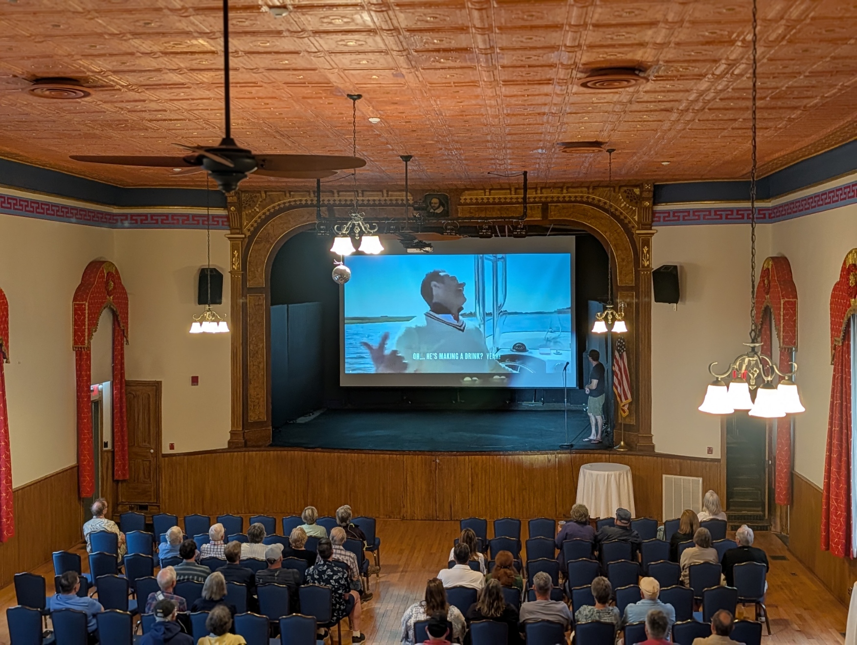 Audience watching a presentation on a large screen in a theater with wooden ceiling and red curtains.