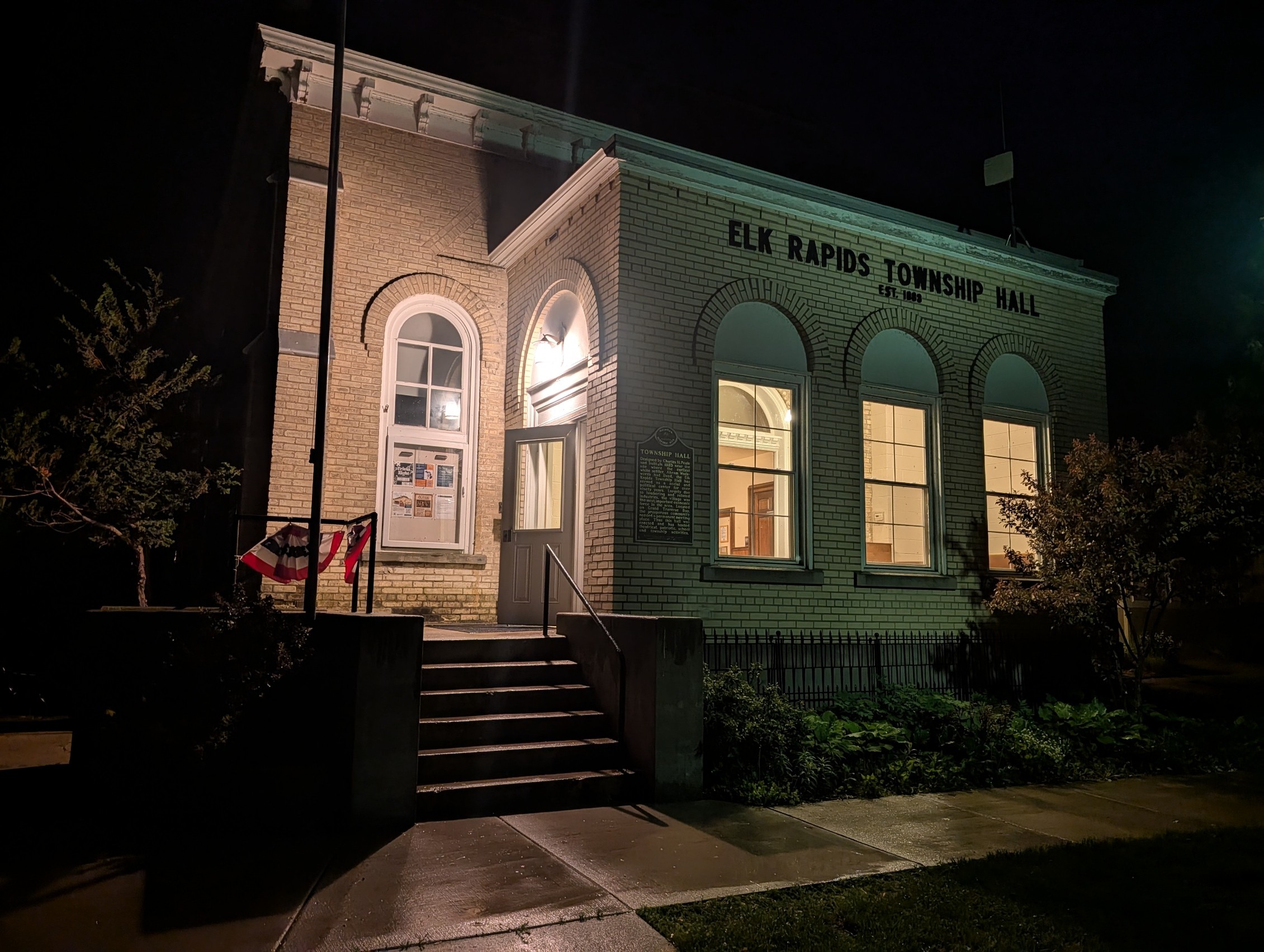 Elk Rapids Township Hall building at night with illuminated windows and entrance, brick exterior, steps, and surrounding greenery.