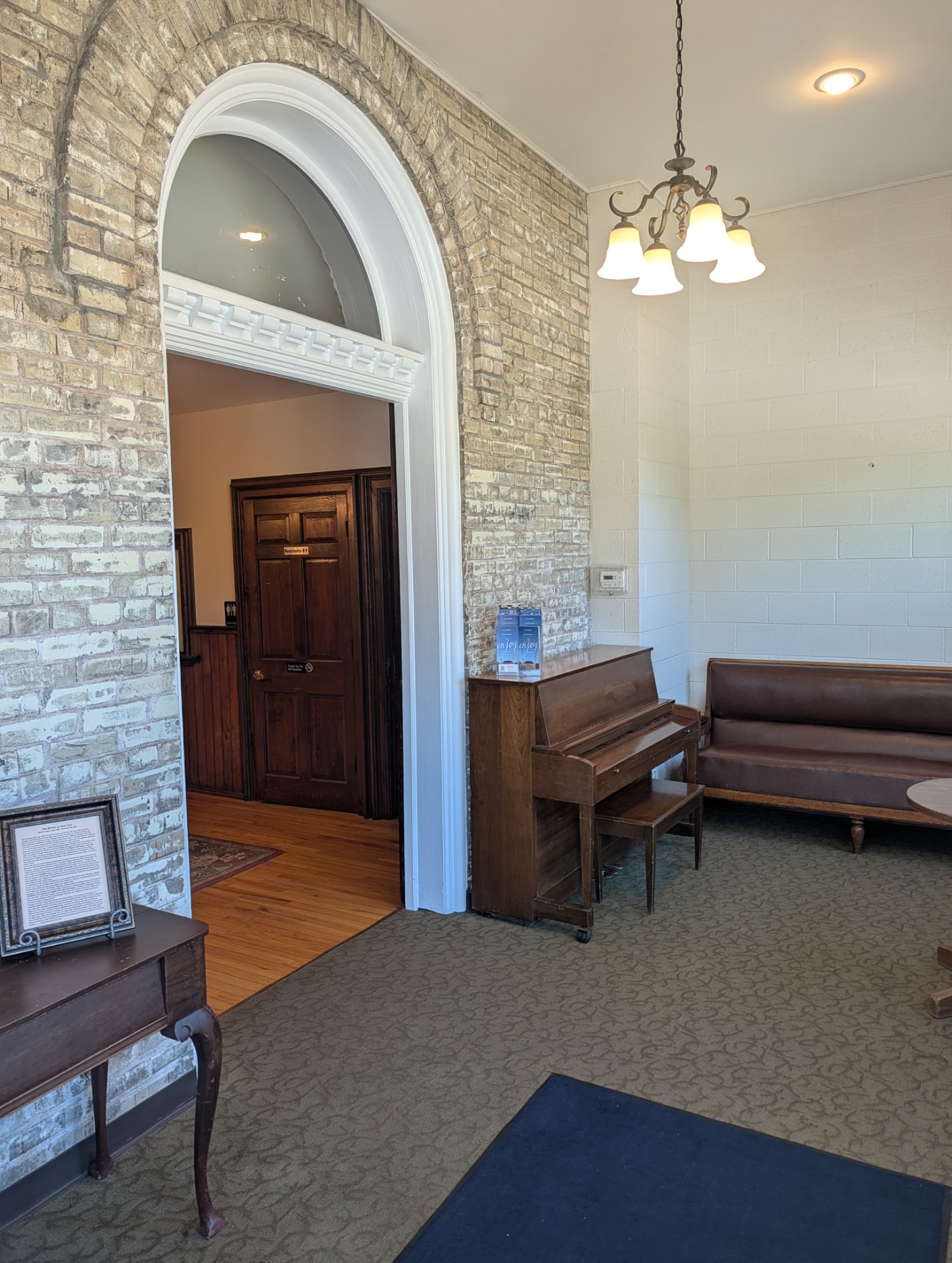 Interior room with brick archway, wooden door, piano, sofa, and carpeted floor, illuminated by ceiling light.