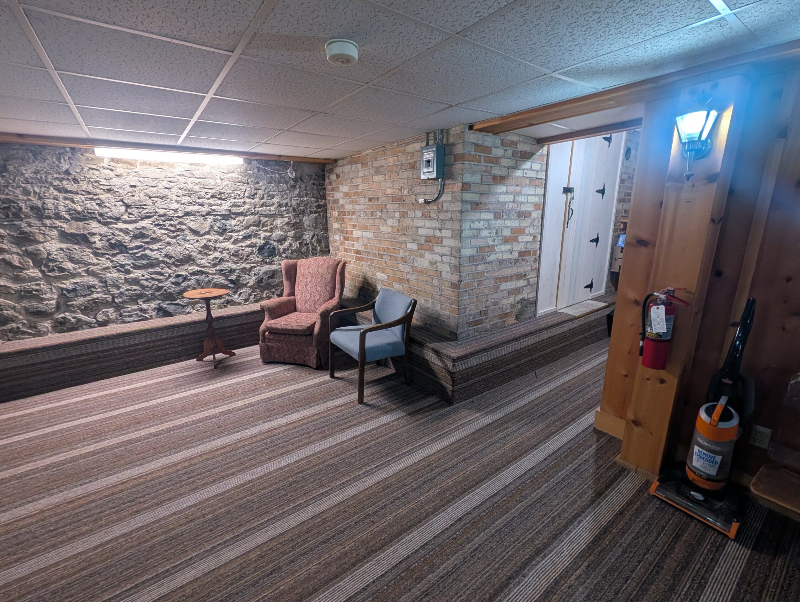 Corner of a room with stone and brick walls, two chairs, a small table, and a striped carpet.