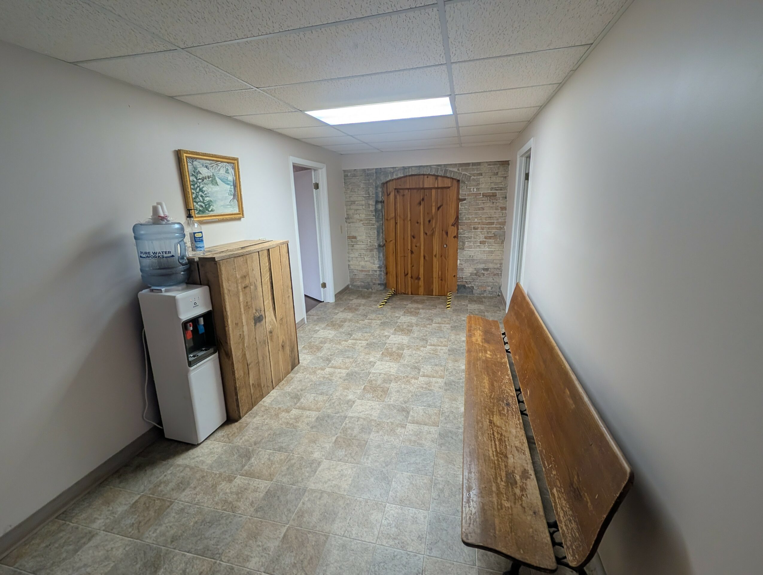 Long narrow hallway with a wooden bench on the right, water cooler on the left, and a wooden door at the end.