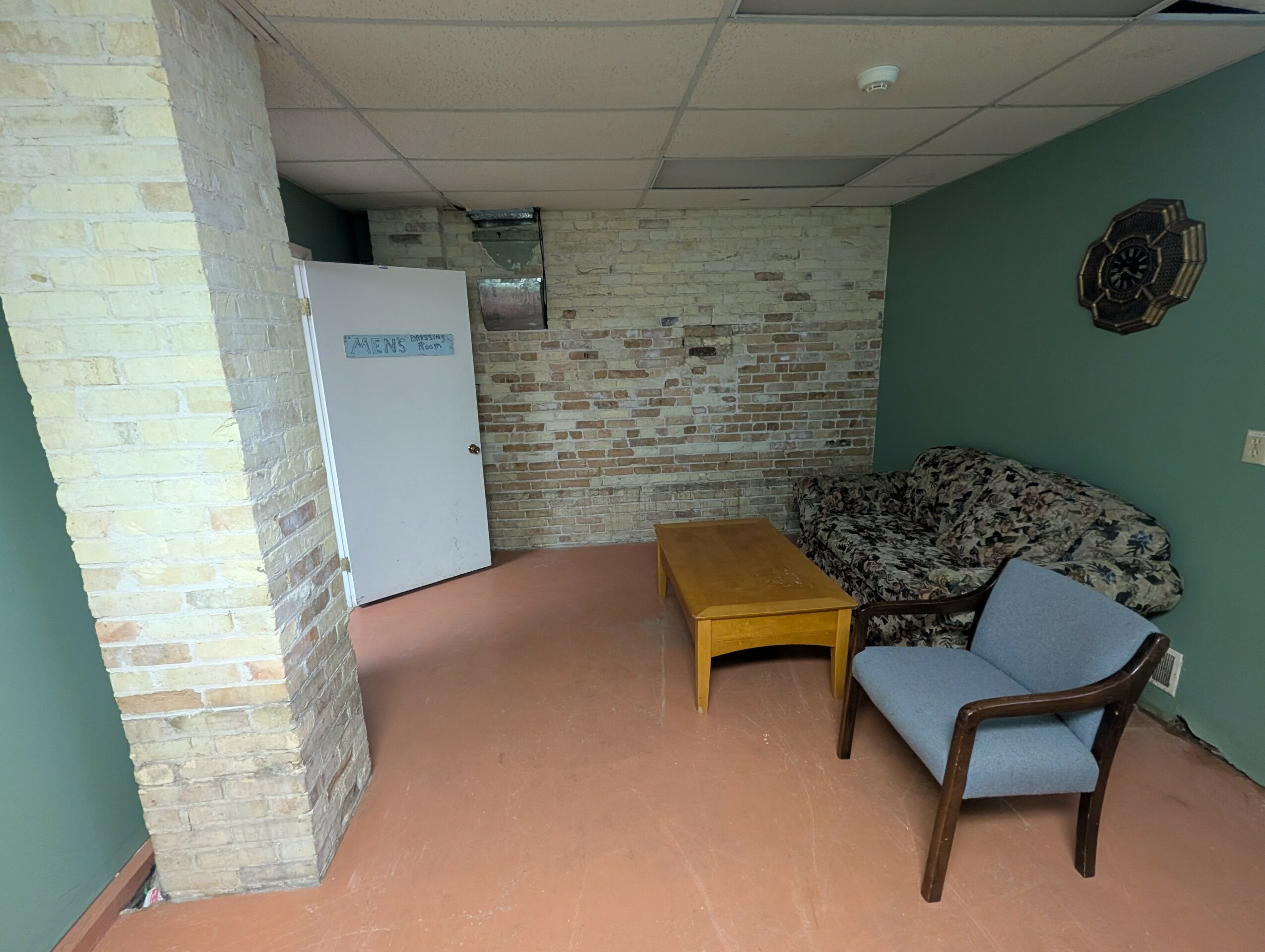 Room with brick wall, sofa, armchair, yellow table, and wall decoration, ceiling with tiles, and a door in the background.
