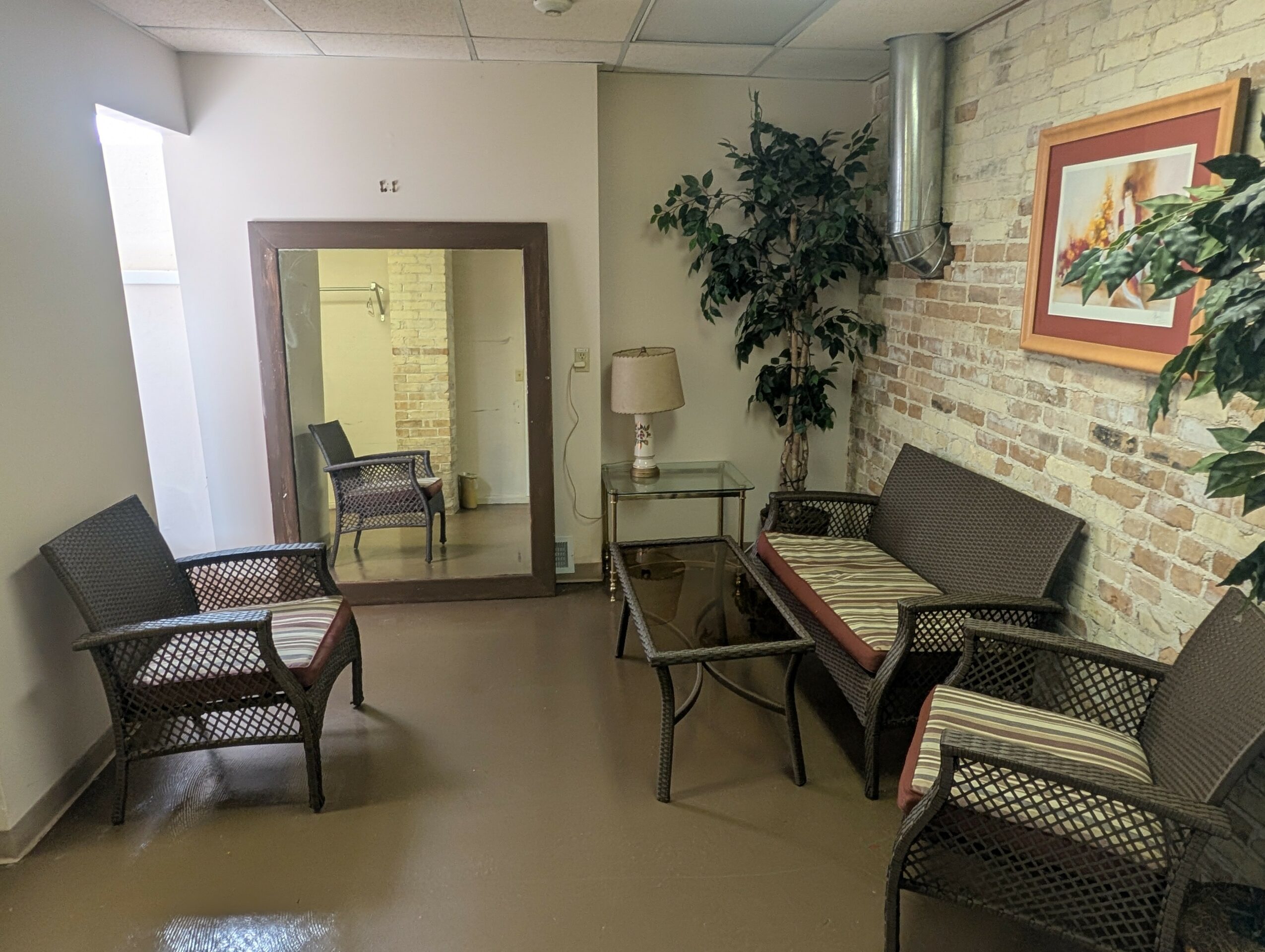 Waiting room with chairs, a glass table, a lamp, a large plant, and framed artwork on a brick wall.