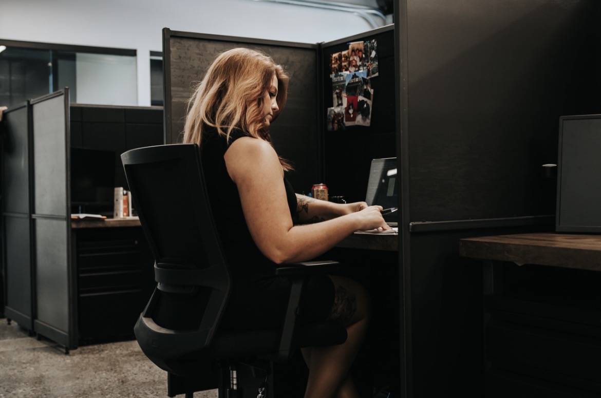 Woman sitting at a desk in an office cubicle, working on a computer, with photos on the wall behind her.