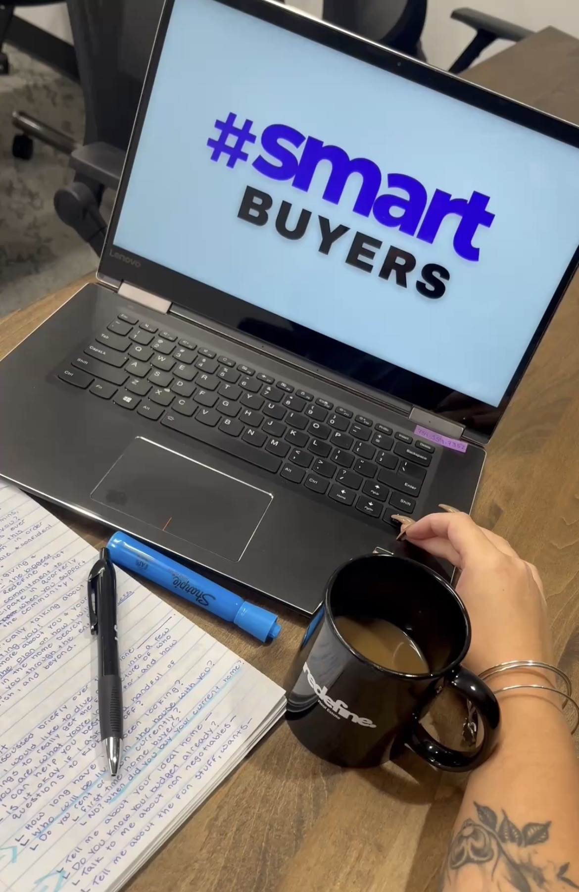 Laptop displaying '#smart BUYERS' on screen, hand holding a mug, notebook, pen, and blue highlighter on table.