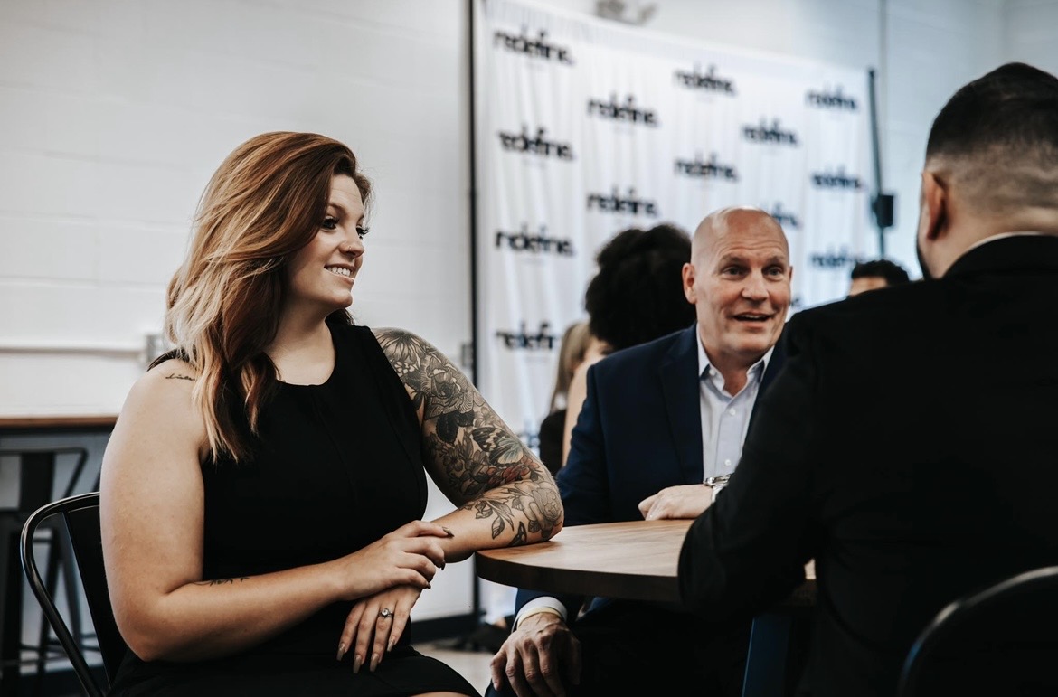 Three people sitting at a table in a meeting room, engaged in conversation, with a backdrop displaying a logo or text.