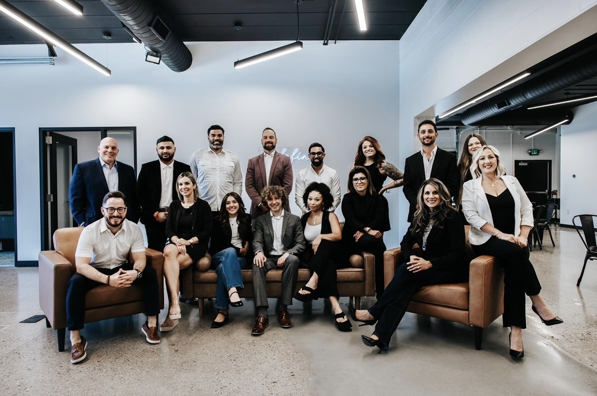 Group of 14 diverse professionals in a modern office, some sitting on sofas, others standing, smiling at the camera.