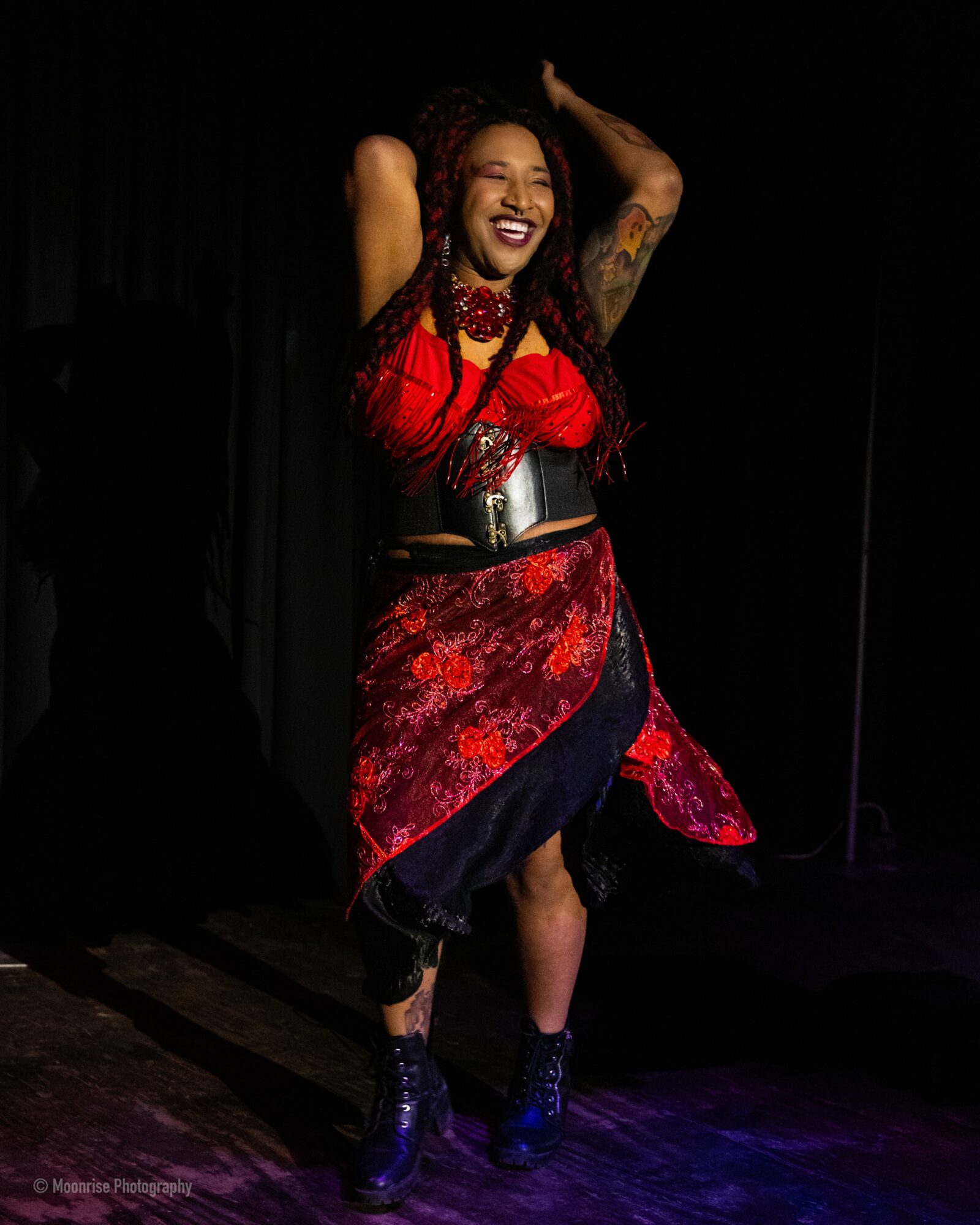 Woman with long hair and tattoos, smiling, wearing a red top, patterned skirt, and blue boots, standing on stage.