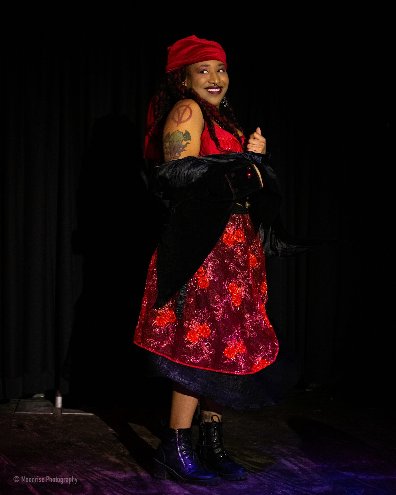 Woman with red headscarf and dress, smiling, standing on stage with black curtain background.