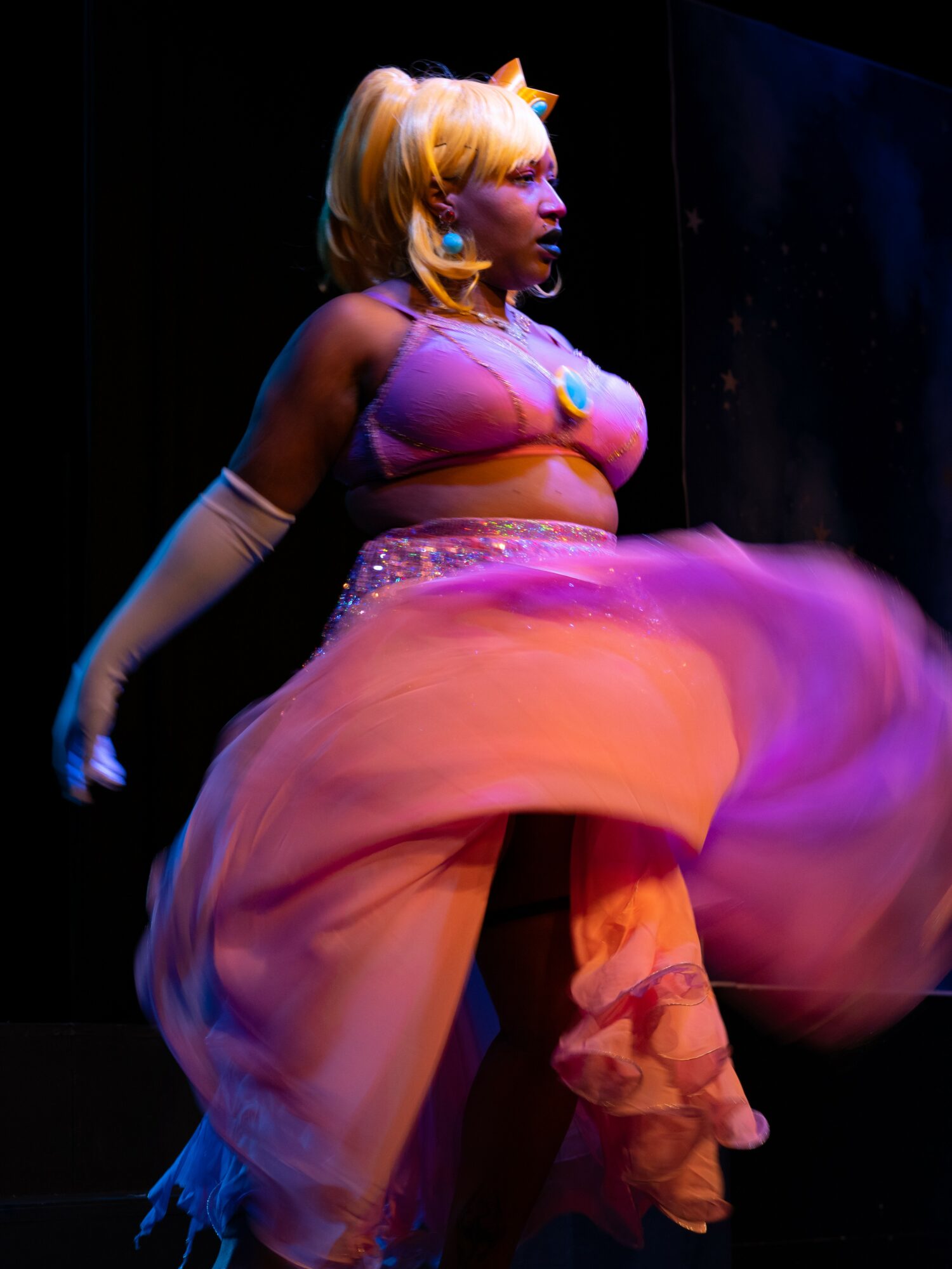 Performer in colorful costume on stage, with a pink and orange flowing skirt and blonde wig, facing sideways.