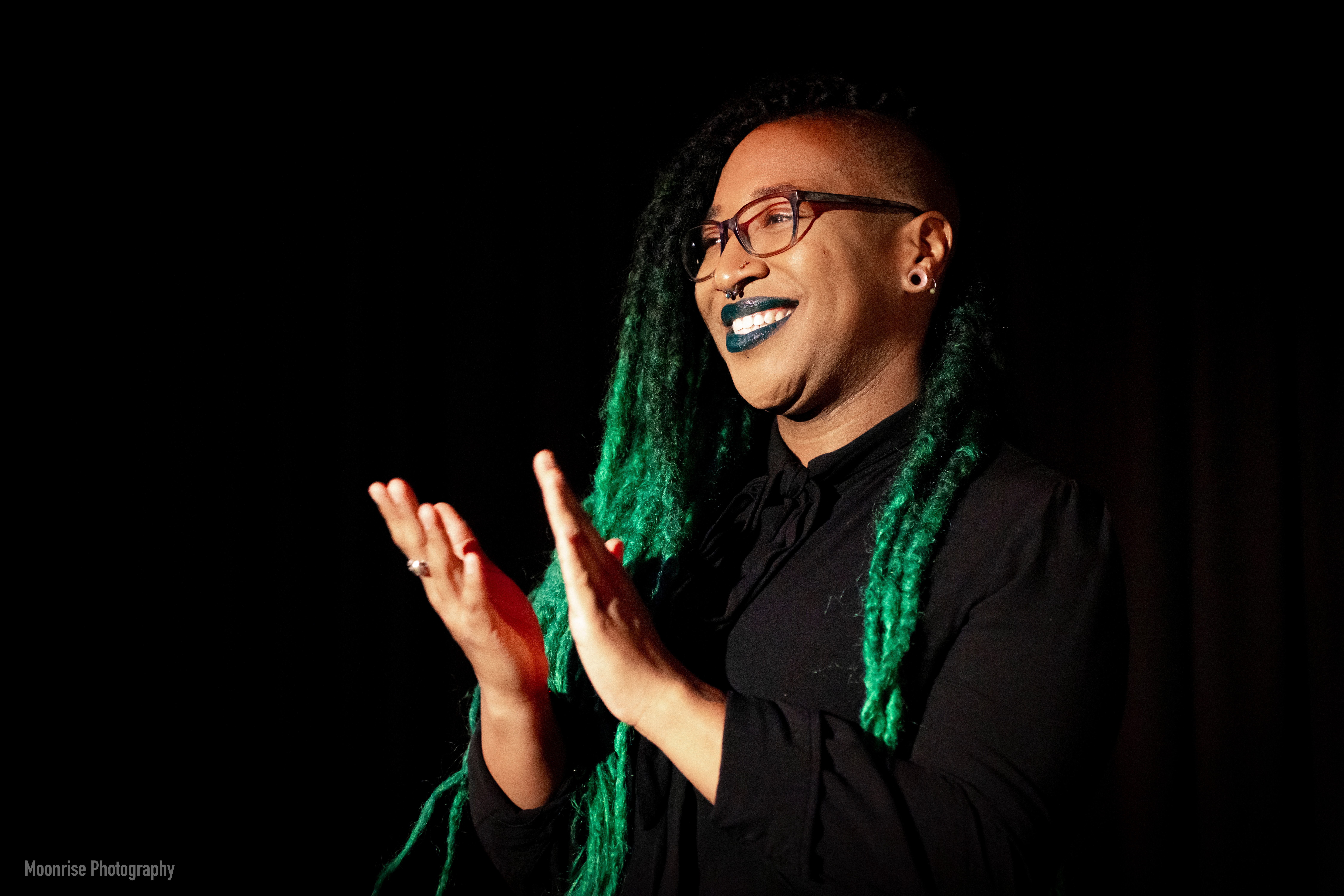 Person with glasses and green braided hair smiling and clapping against a black background.
