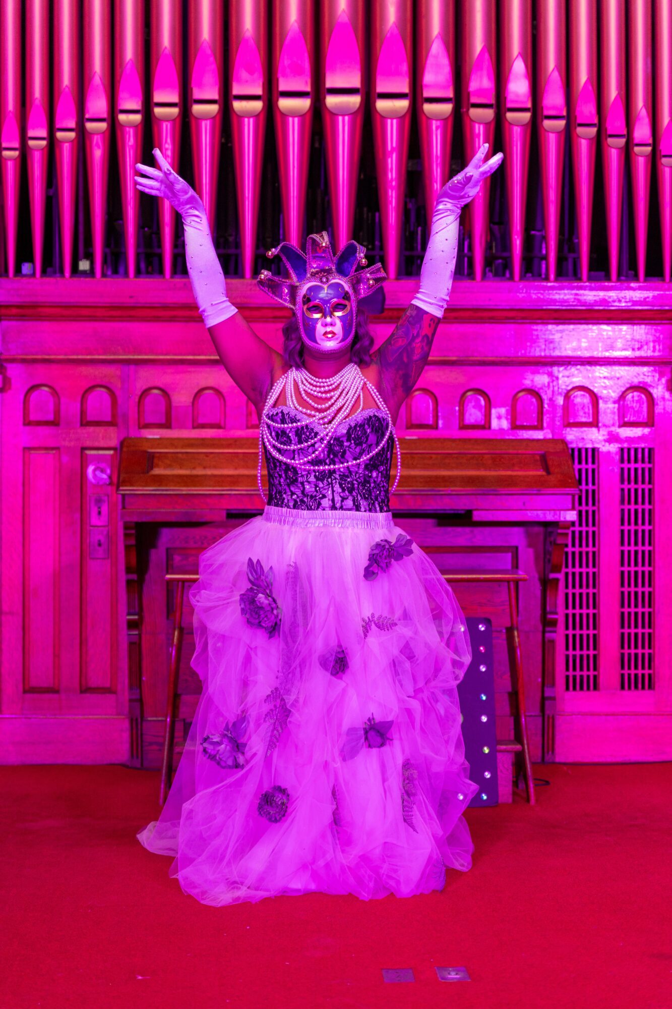 Person in a colorful costume with a mask, standing with arms raised in front of a pipe organ, pink lighting.
