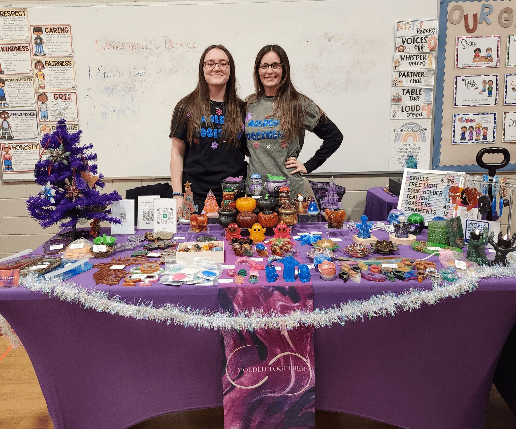 Two women stand behind a decorated table with colorful crafts and ornaments, in a classroom setting. Word posters are on the wall.