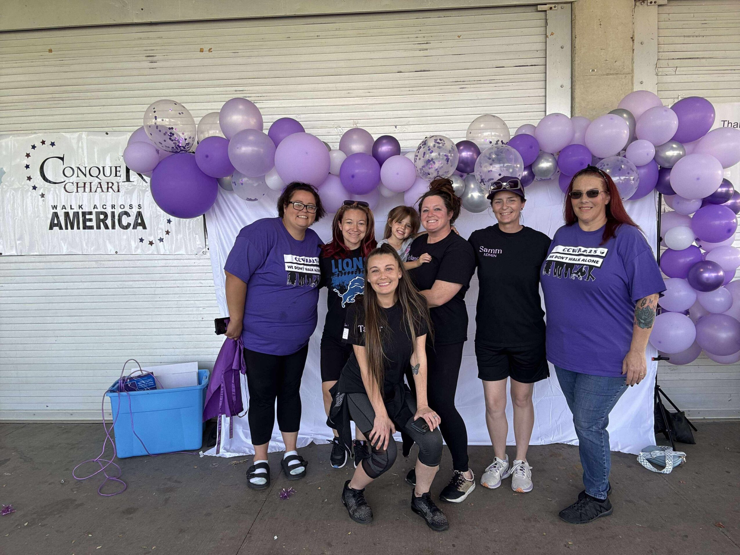 Group of seven people standing in front of purple and white balloon arch, smiling, with a closed shutter background.