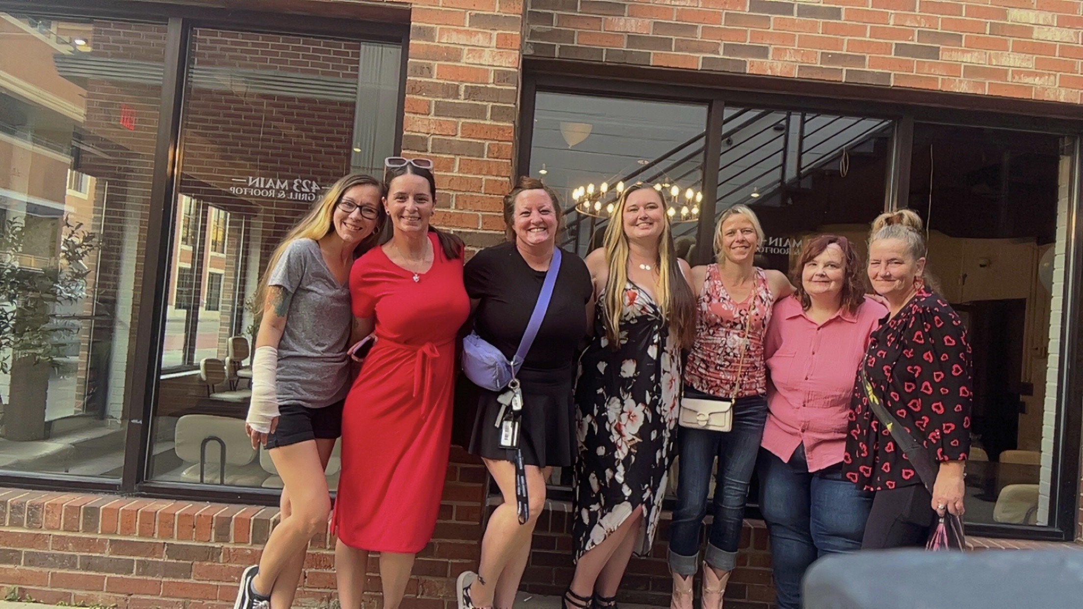 Seven women standing outside a brick building, smiling, with large windows behind them.