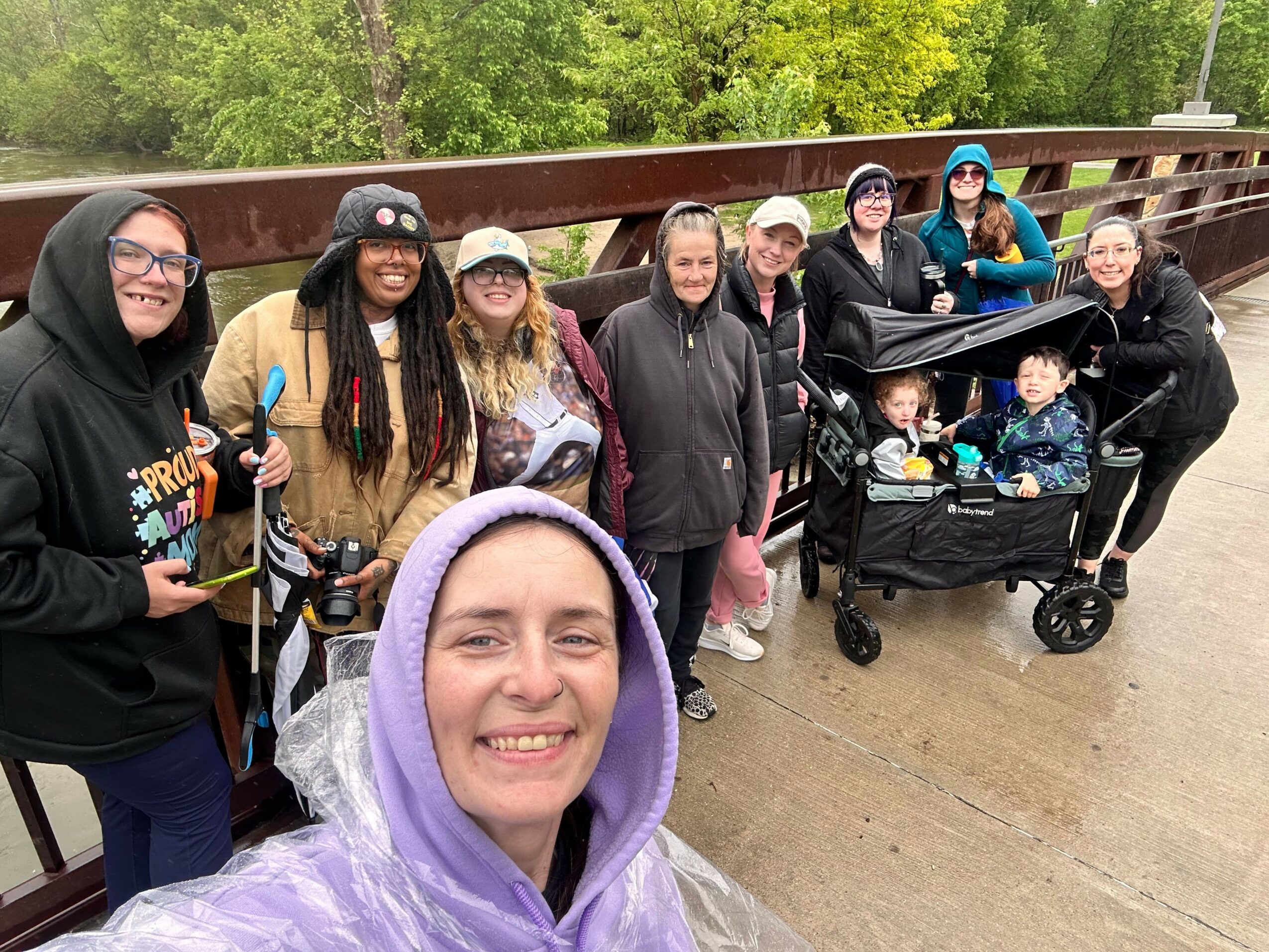 Group of people outdoors, some wearing masks, standing on a bridge with green trees in the background.