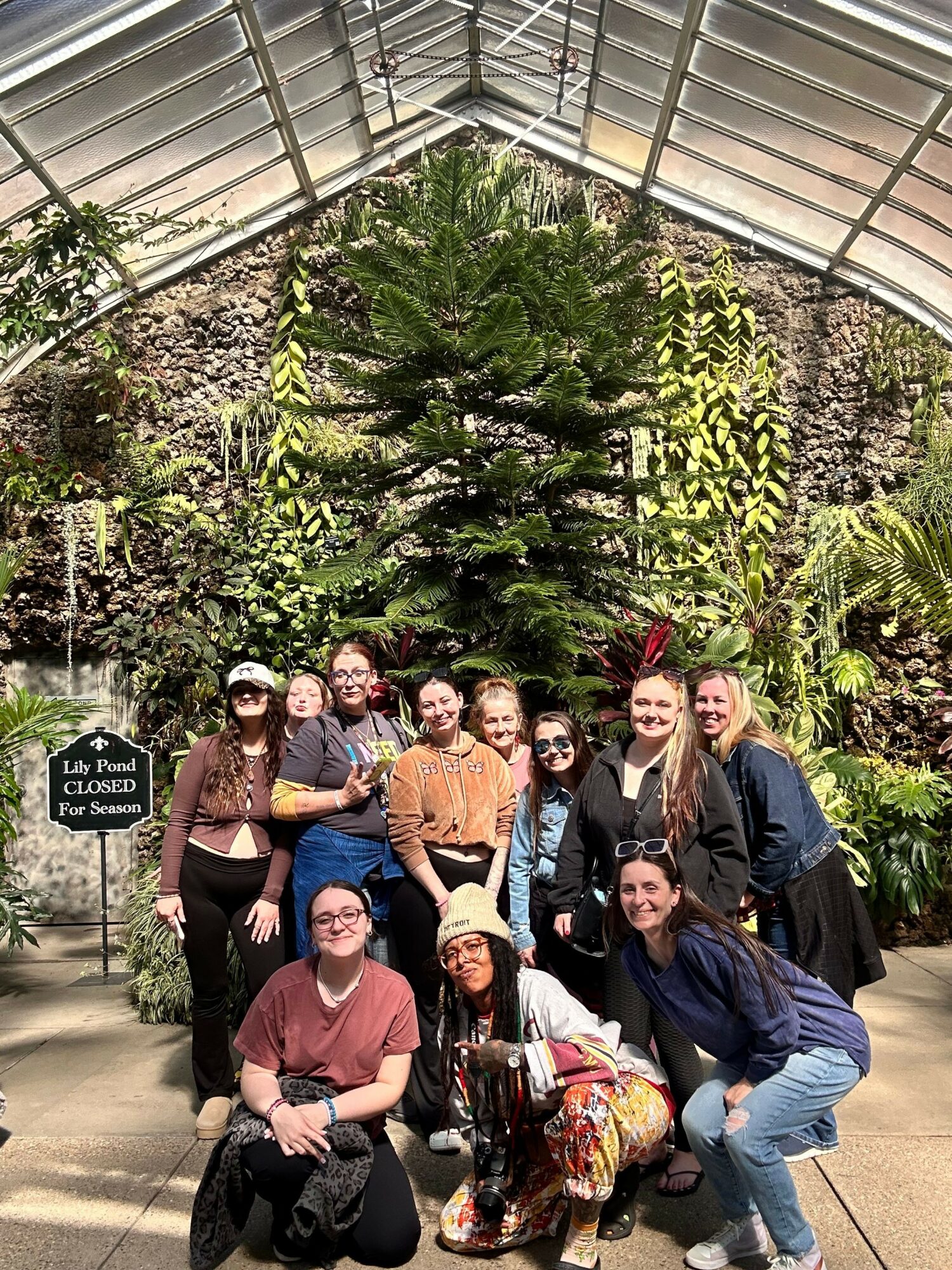 Group of eight people standing outdoors in a garden area with plants and trees, some holding drinks, smiling, and posing for the photo.