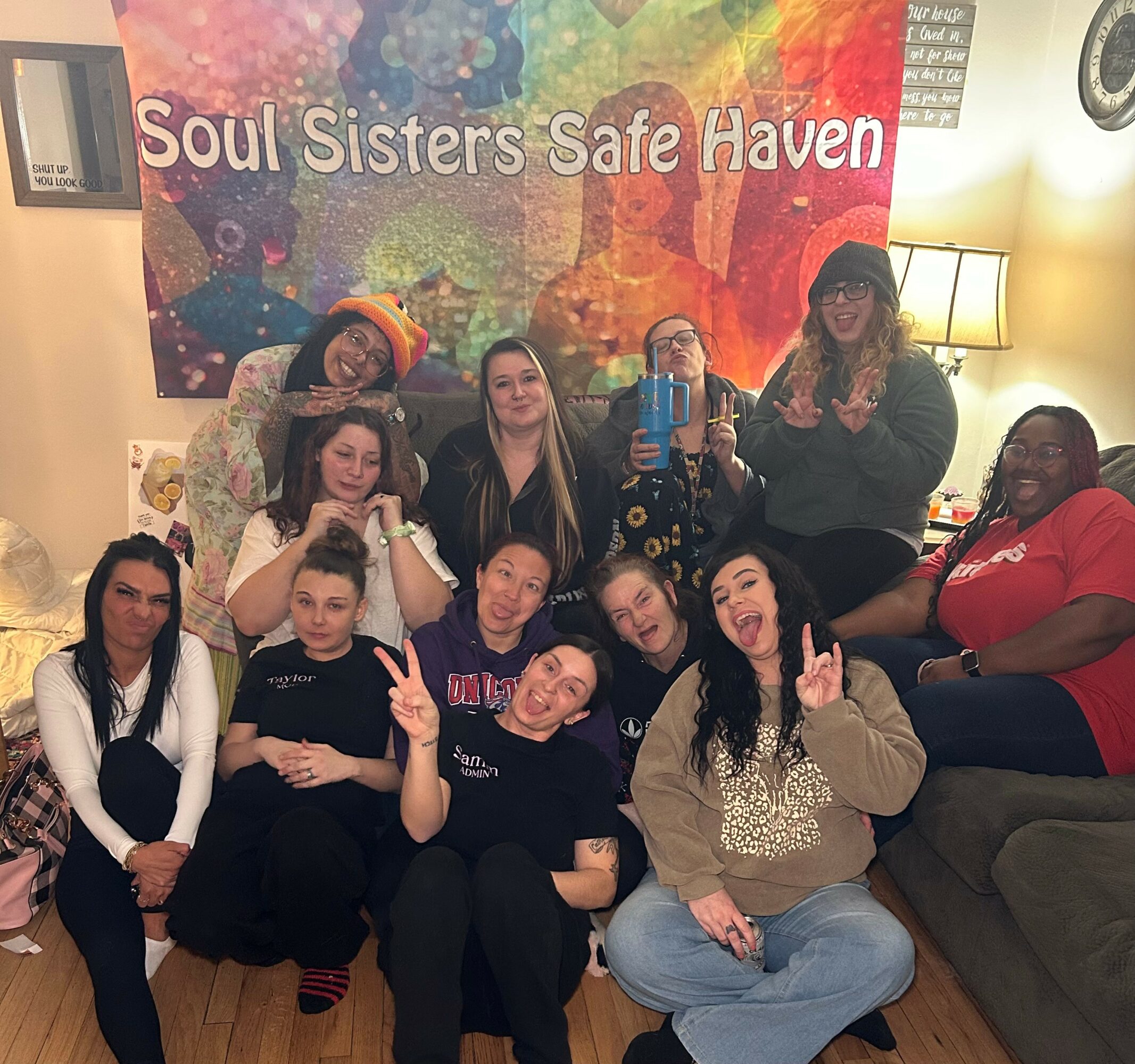 Group of women and girls sitting and standing in a living room with a colorful banner reading 'Soul Sisters Safe Haven' behind them.