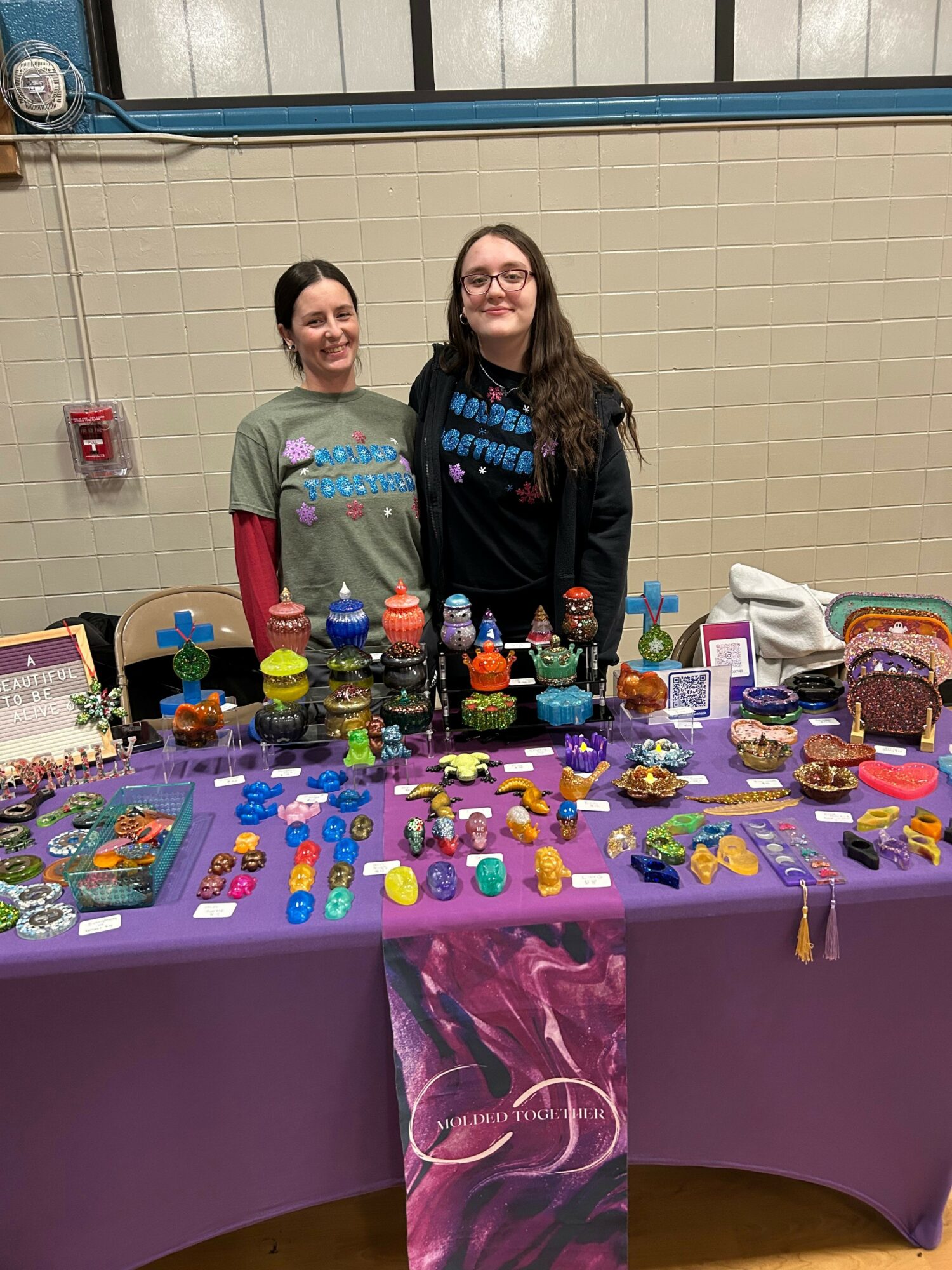 Two women stand behind a table filled with colorful toys and items, in an indoor setting with a beige wall.