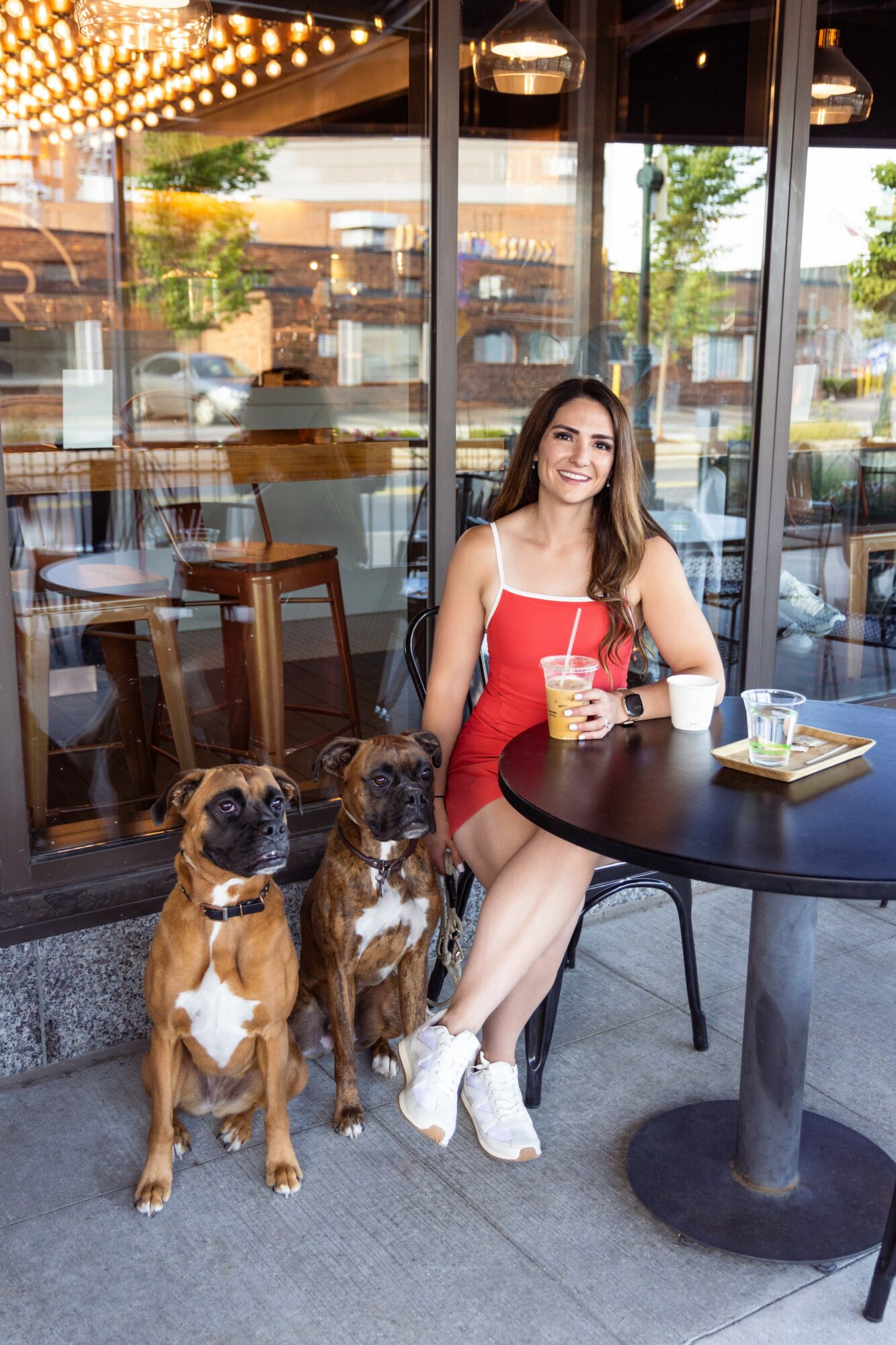 Woman in red dress sitting at outdoor cafe table with two dogs, holding a drink, smiling, glass windows behind.