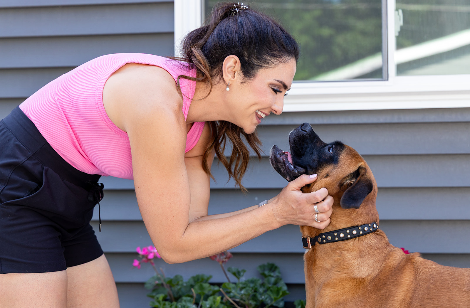 Woman in pink top and black shorts playing with a dog outside near a house wall.