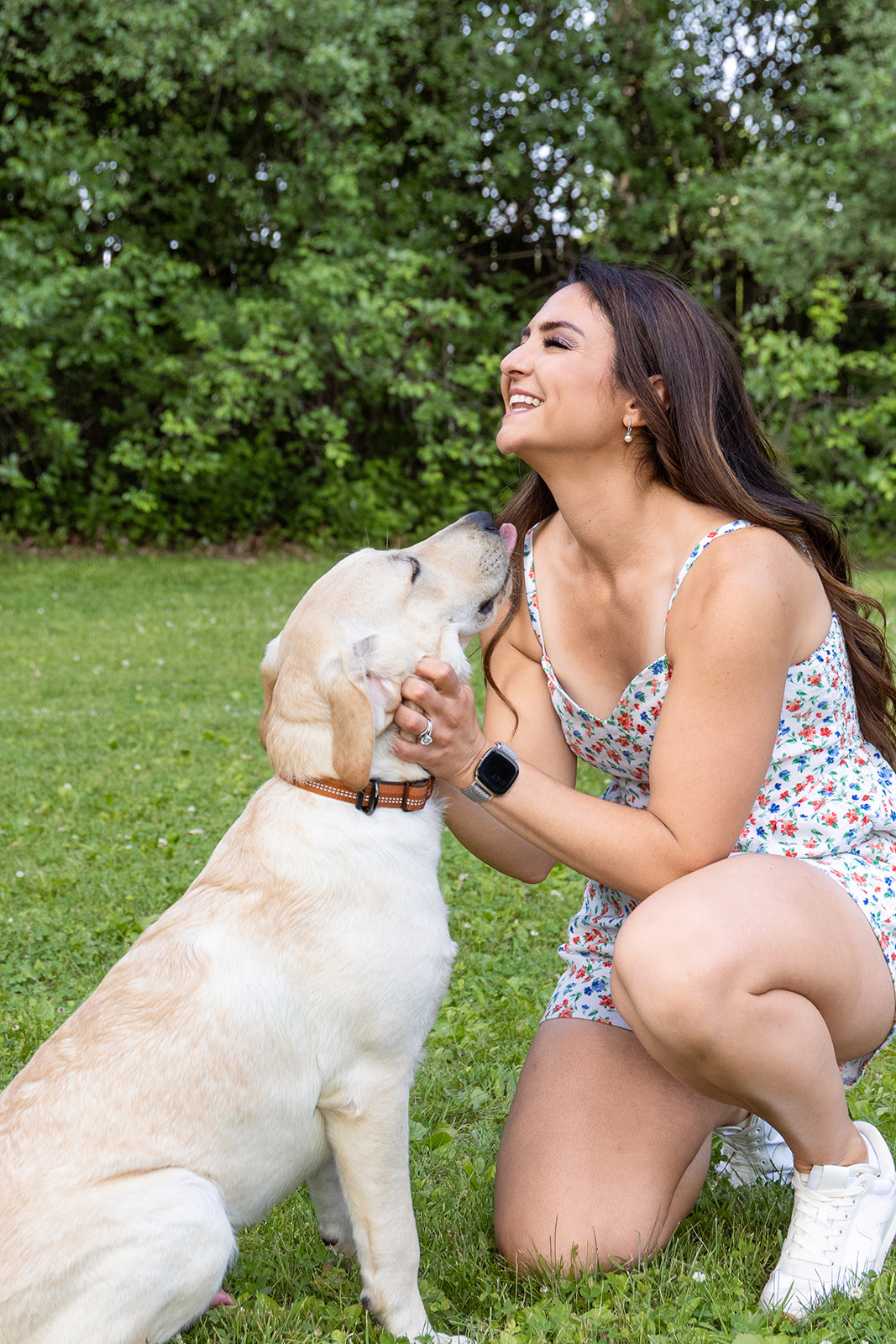 Young woman kneeling on grass, smiling, with dog licking her face, surrounded by green trees.