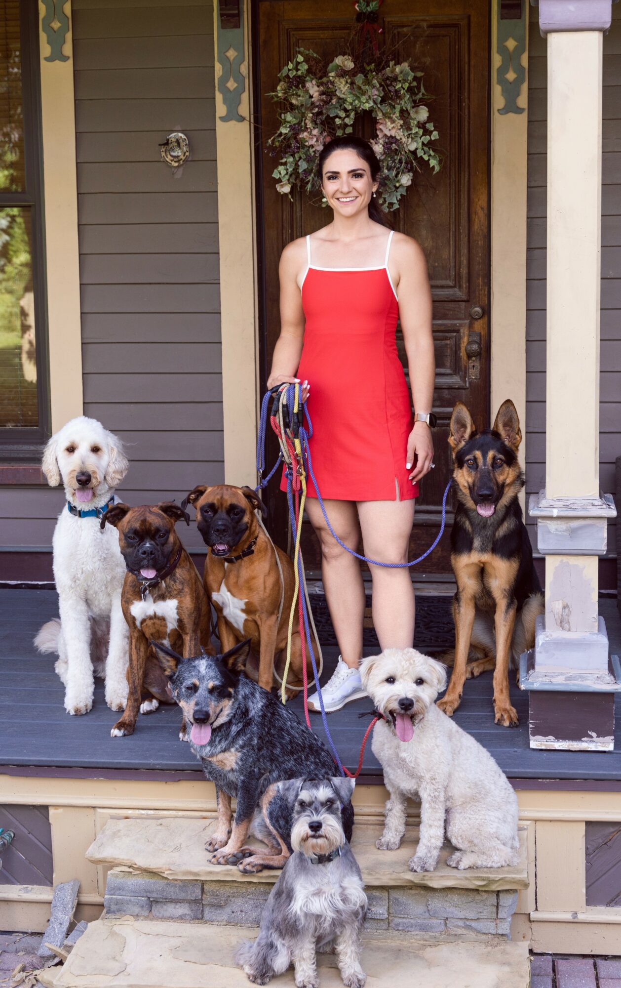 Woman in red dress standing on porch with five dogs, house door and floral wreath in background.