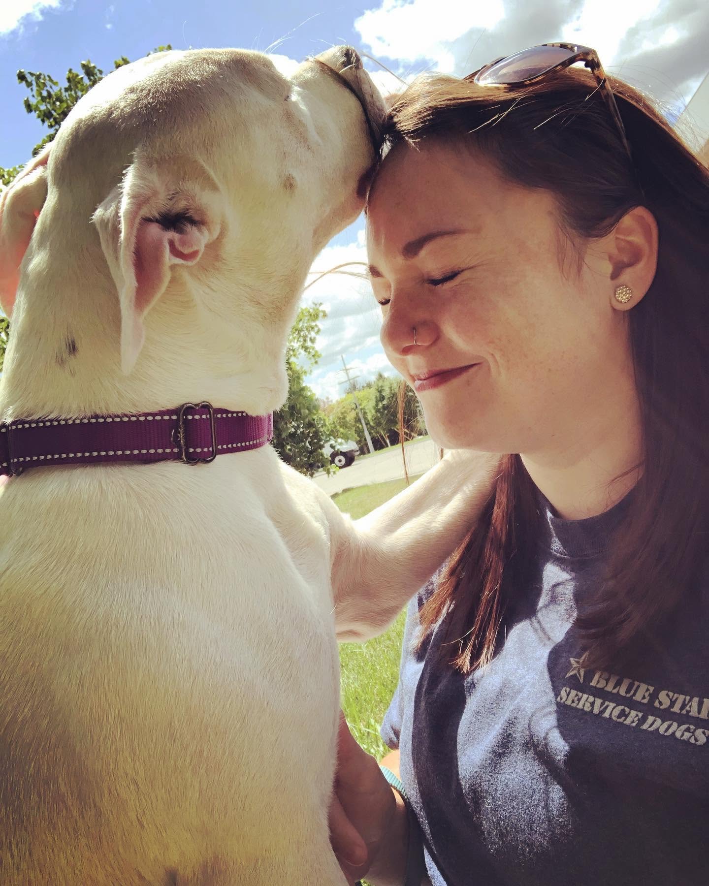 Person with closed eyes smiling as a dog licks their forehead outdoors on a sunny day.