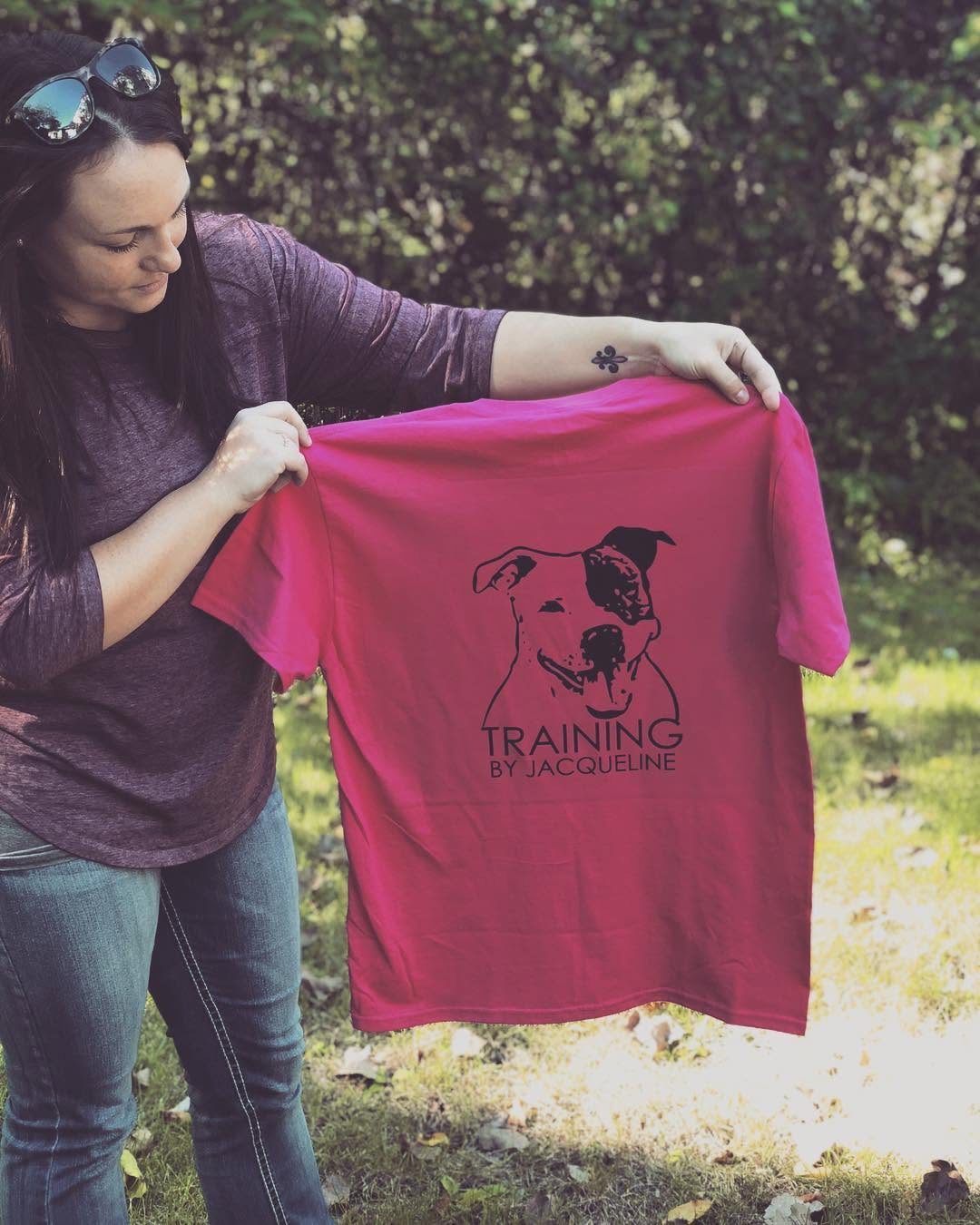 Person holding a pink T-shirt with a dog illustration and text, outdoors with trees and grass in background.