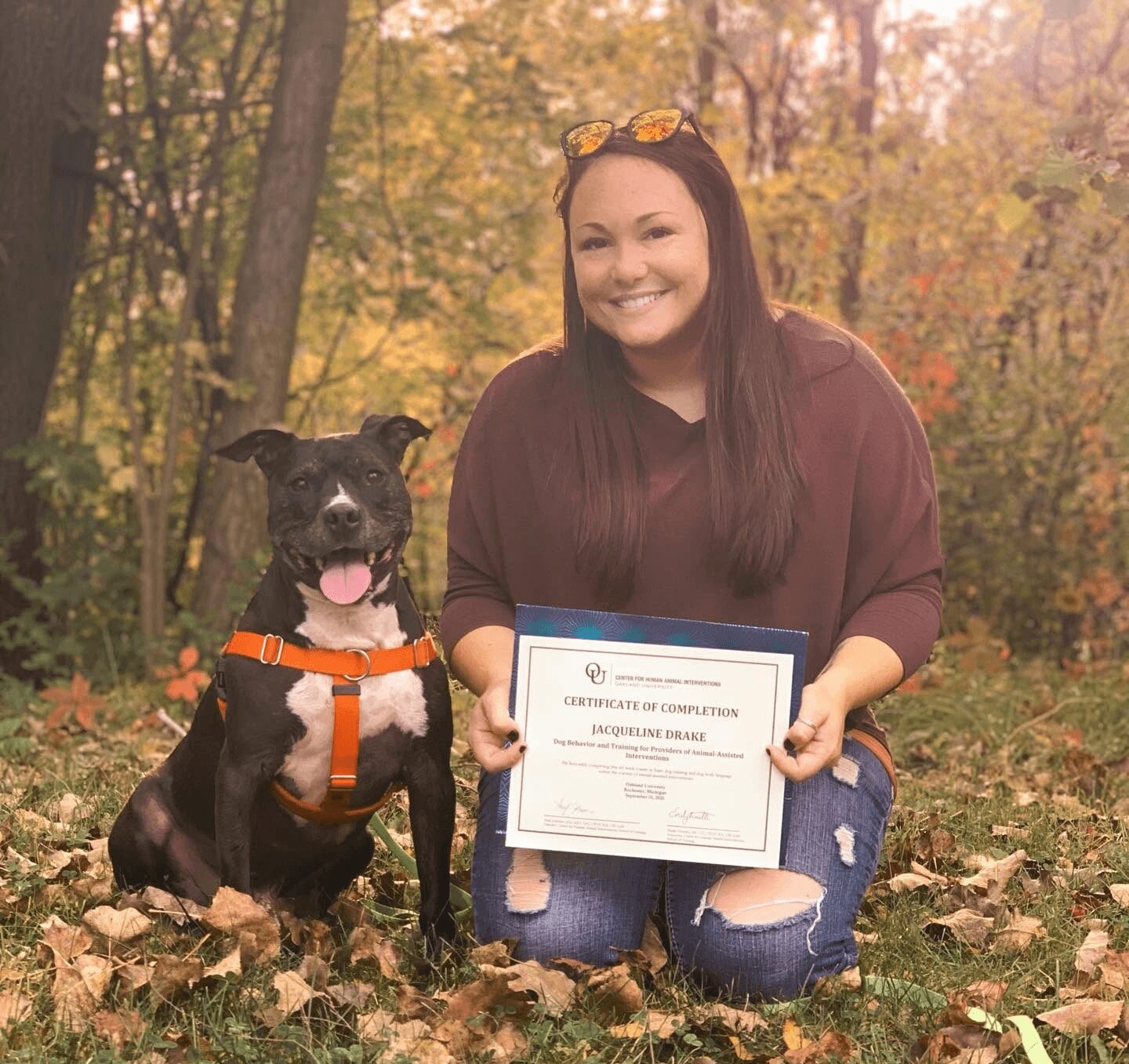 Woman kneeling outdoors with a dog holding a certificate, surrounded by autumn leaves and trees.