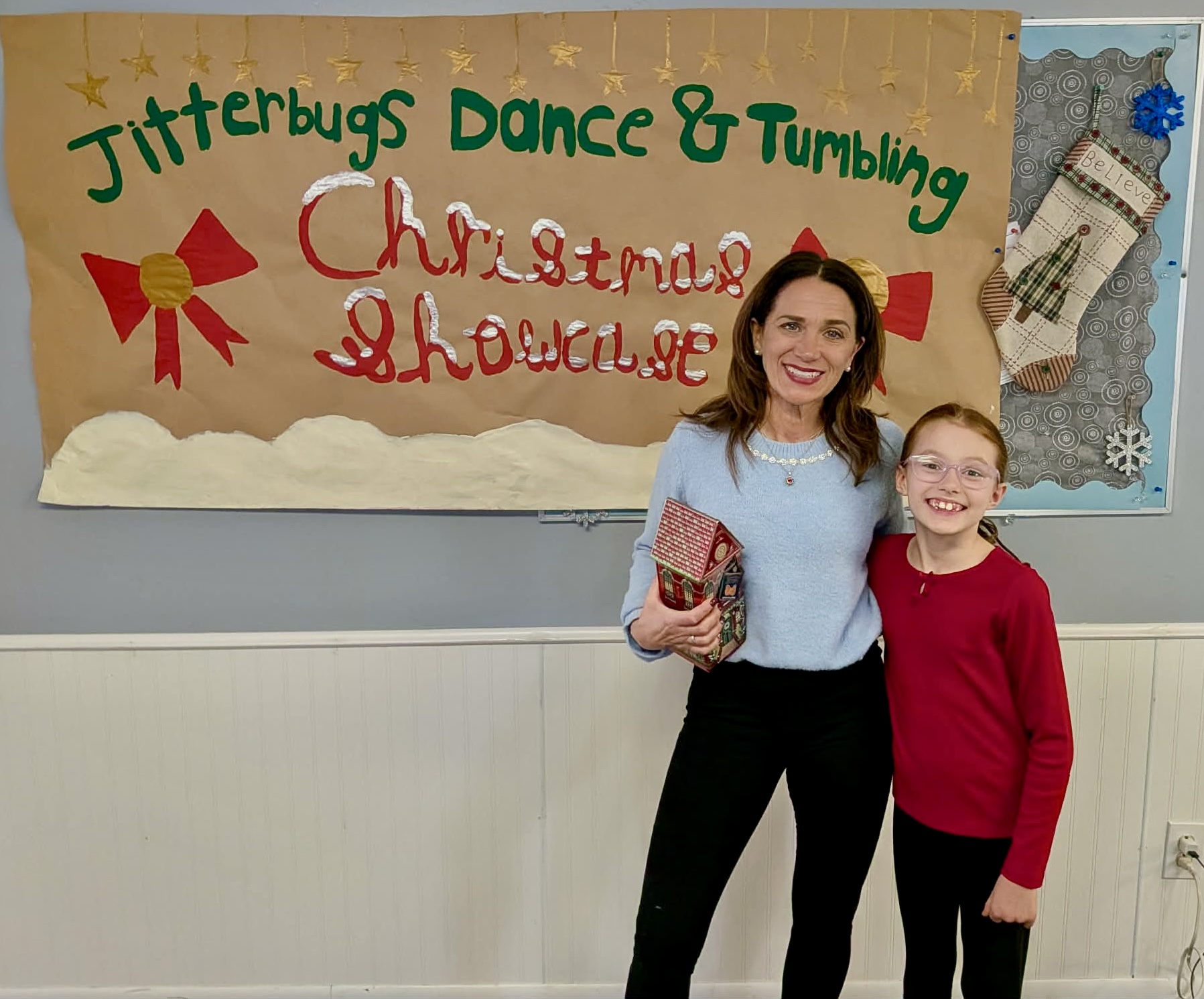 Two smiling females, one adult and one child, stand in front of a decorated bulletin board with holiday-themed decorations and text.