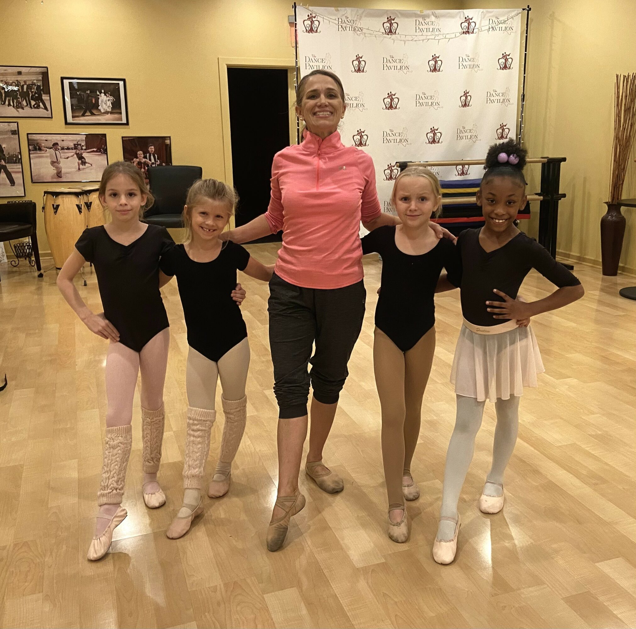 Group of four young girls in dance attire with an instructor in a dance studio, smiling and posing together.