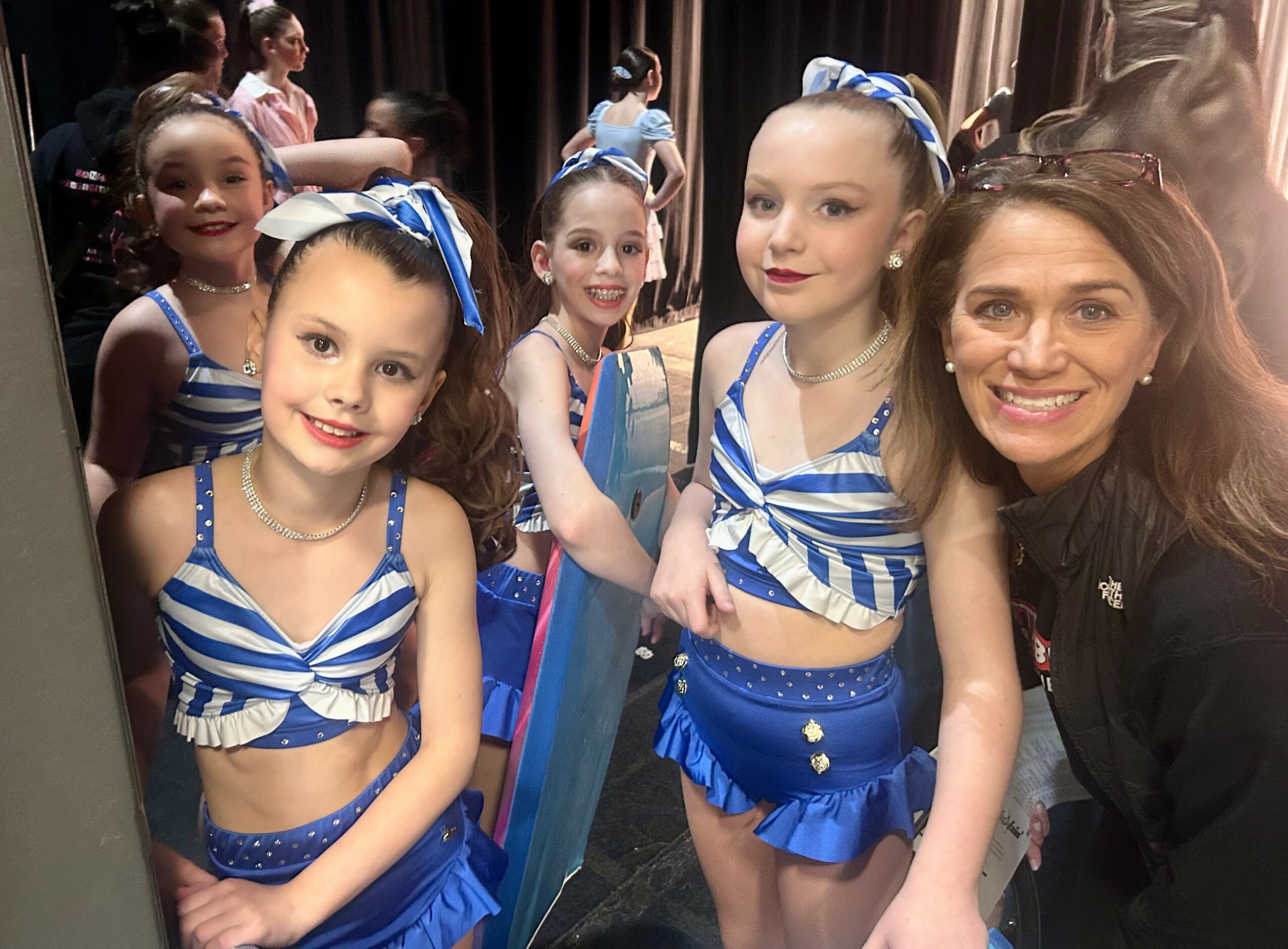 Group of young girls in blue and white costumes with a woman, smiling backstage, some girls with blue bows in hair.