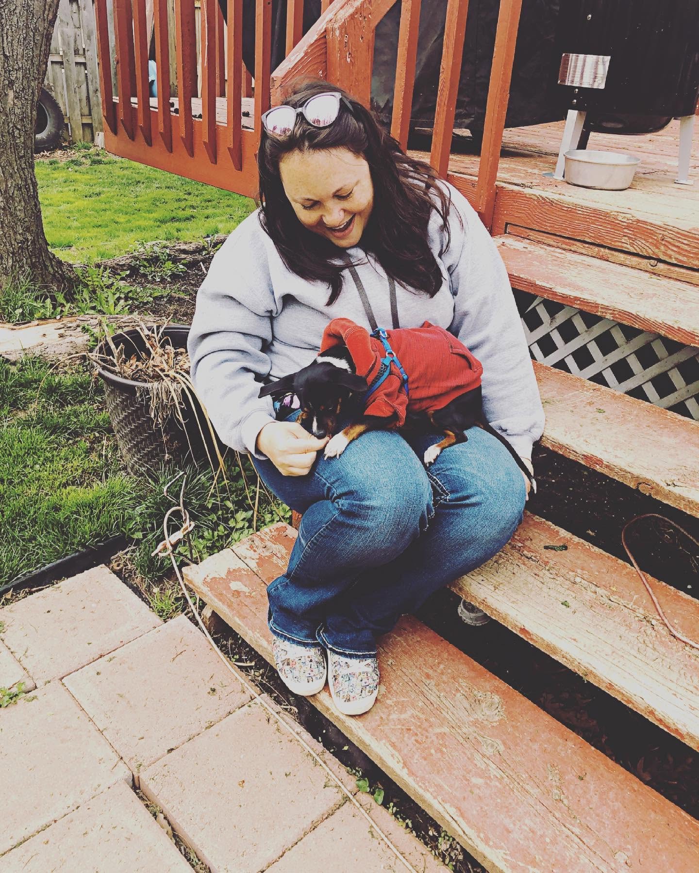 Woman sitting on steps holding a small dog wearing a red jacket outdoors, with a grassy yard and wooden deck in background.