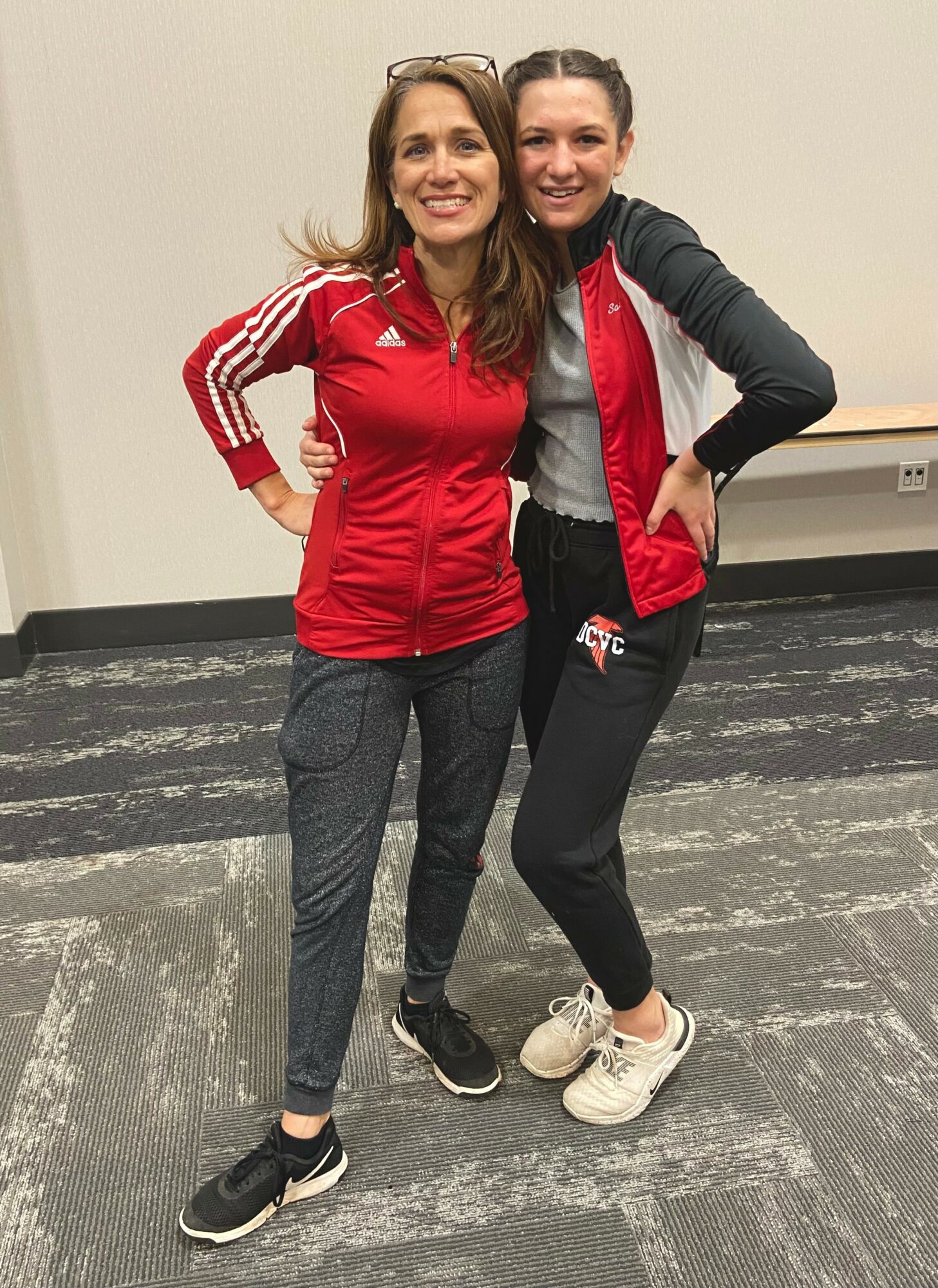 Two women standing together, smiling, with arms around each other, in casual athletic clothing, against a plain wall background.