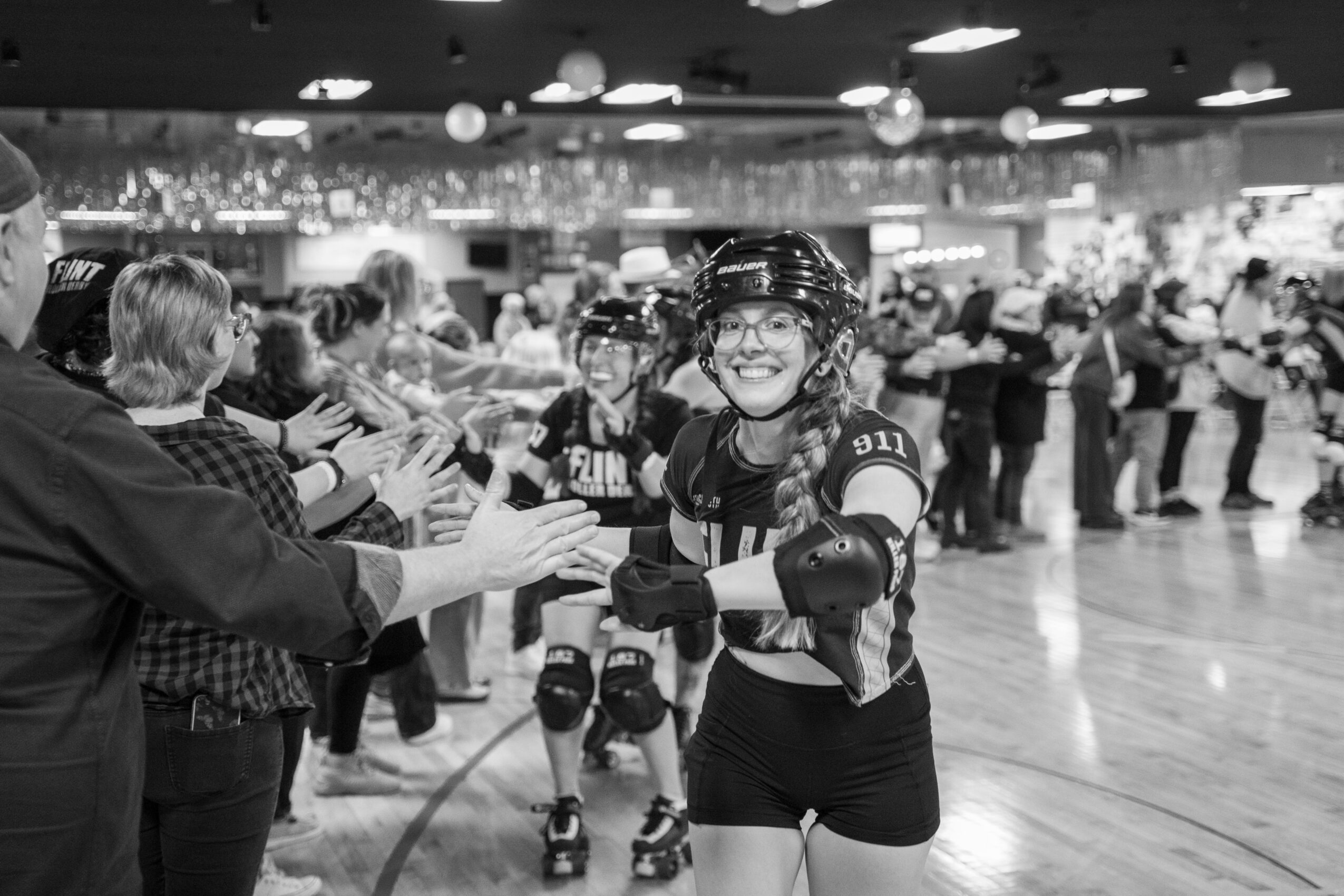 People at roller skating rink greeting and high-fiving, with smiling girl in helmet and protective gear in foreground.