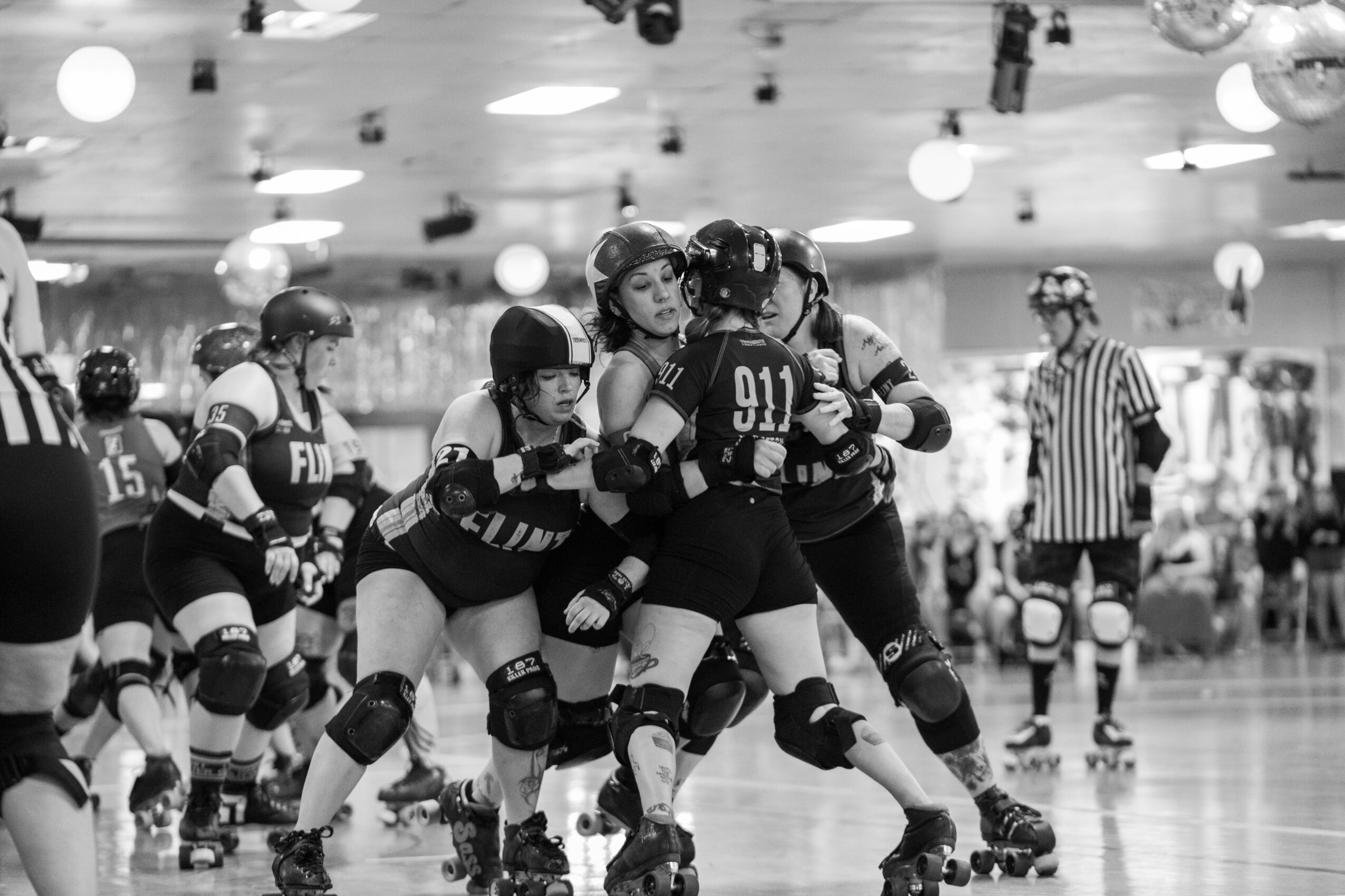Group of roller derby players in protective gear, celebrating together indoors, with a referee in the background.