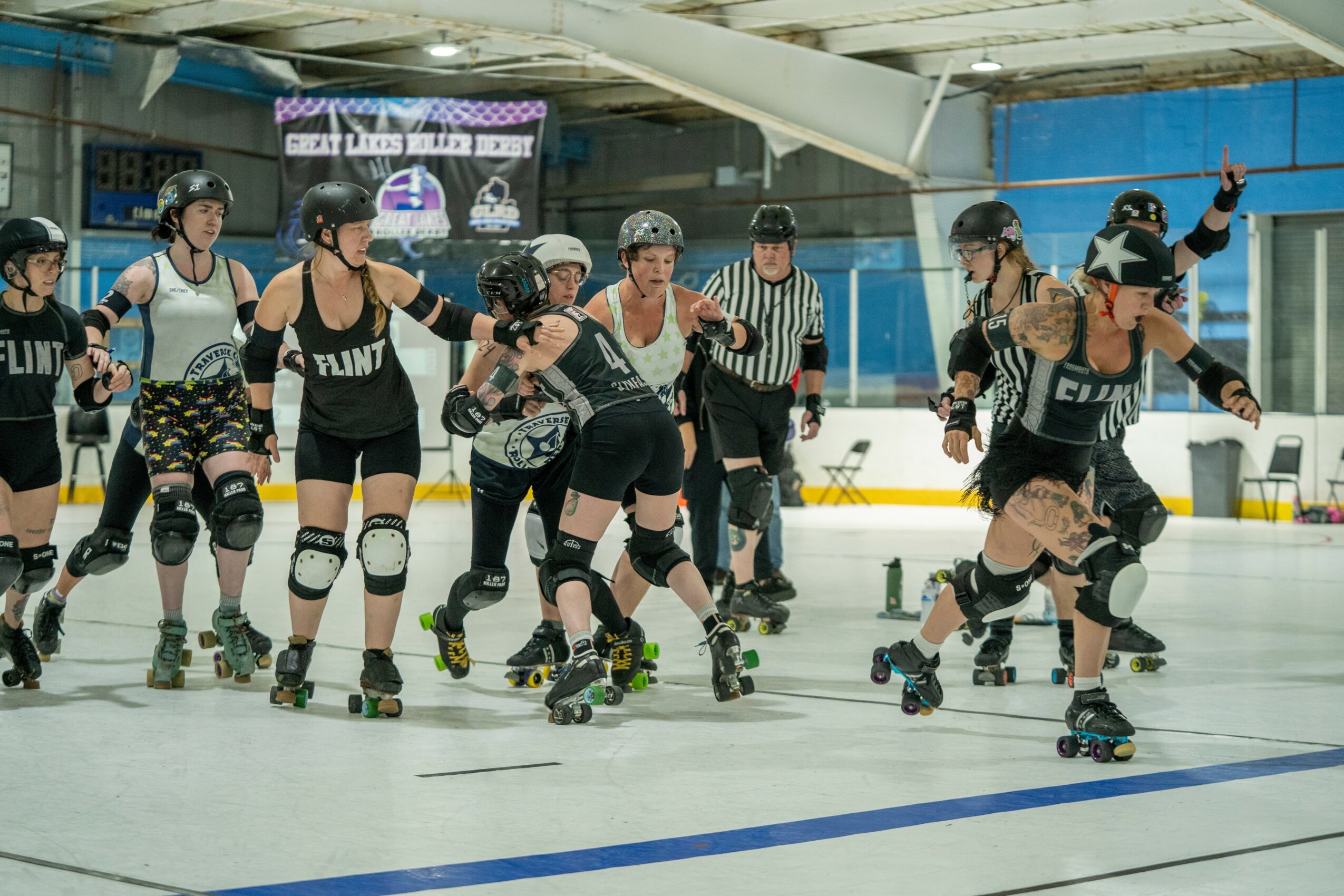 Group of roller derby players in protective gear on an indoor rink, some skating and others standing, with a referee nearby.