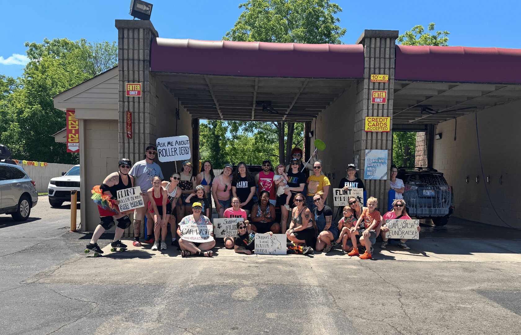 Group of people holding signs in front of a building with cars parked nearby, trees in the background.