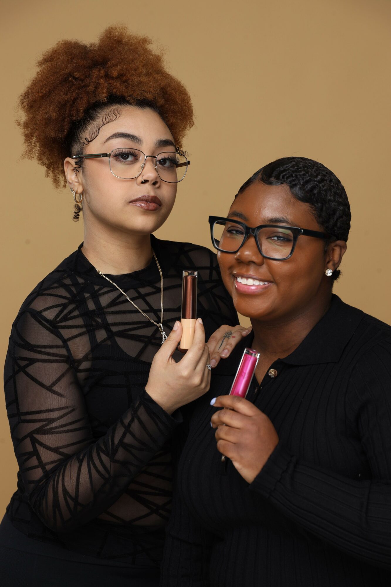 Two women with glasses, one with curly hair and the other with braided hair, smiling and holding makeup brushes.