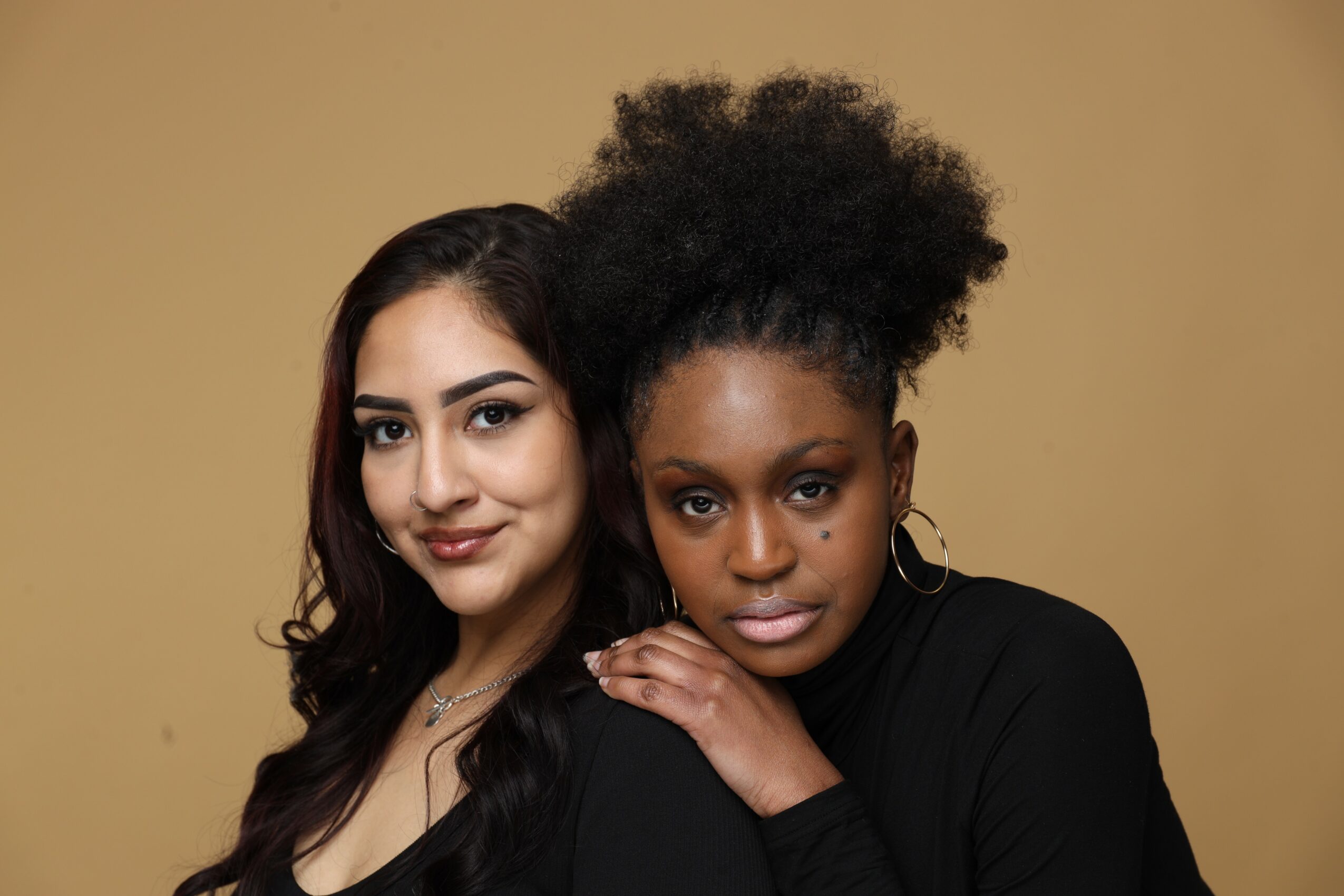 Two women pose closely, one with long dark hair and the other with curly hair, against a beige background.