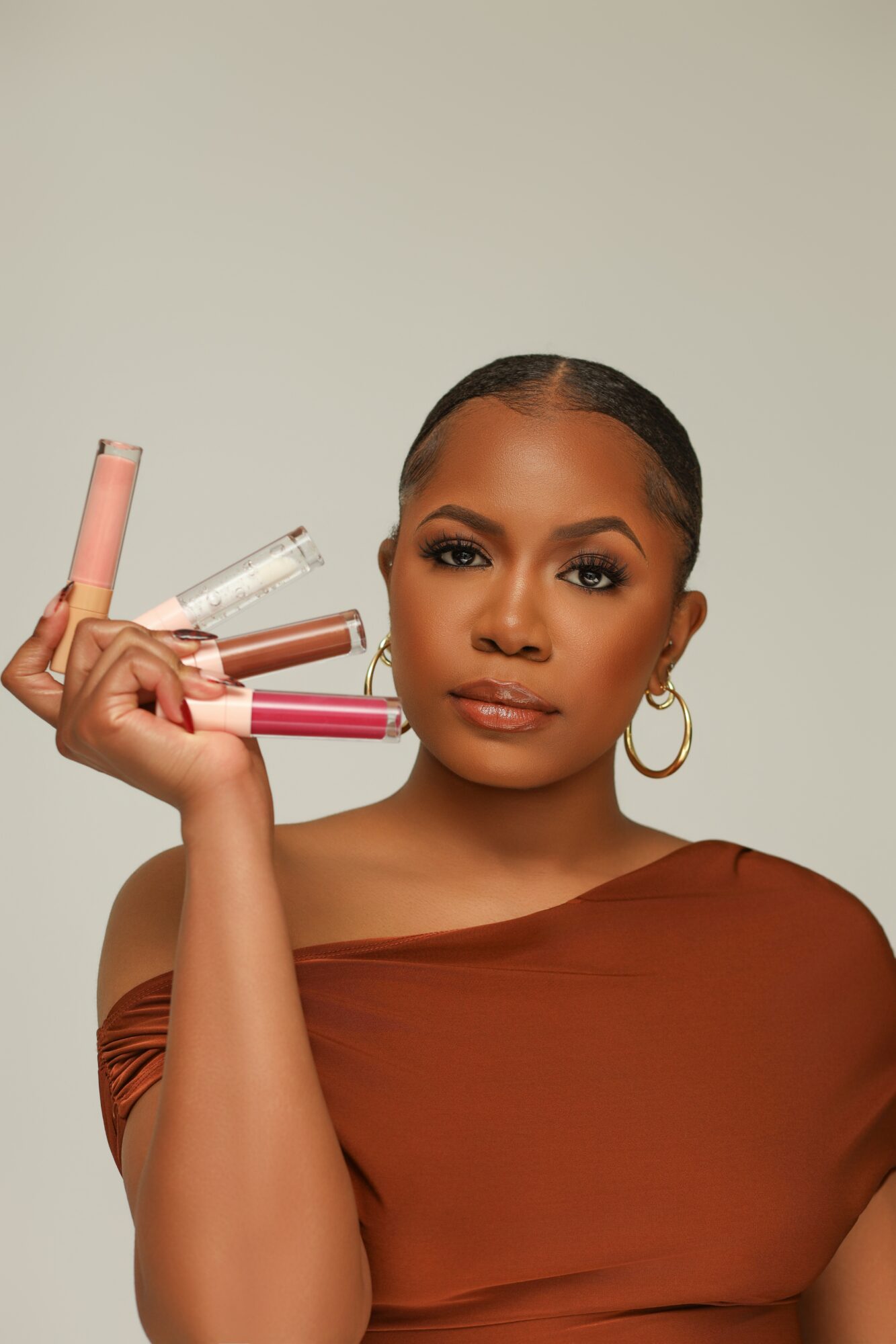 A woman with dark hair and hoop earrings holds several lip gloss tubes near her face, wearing a brown top.