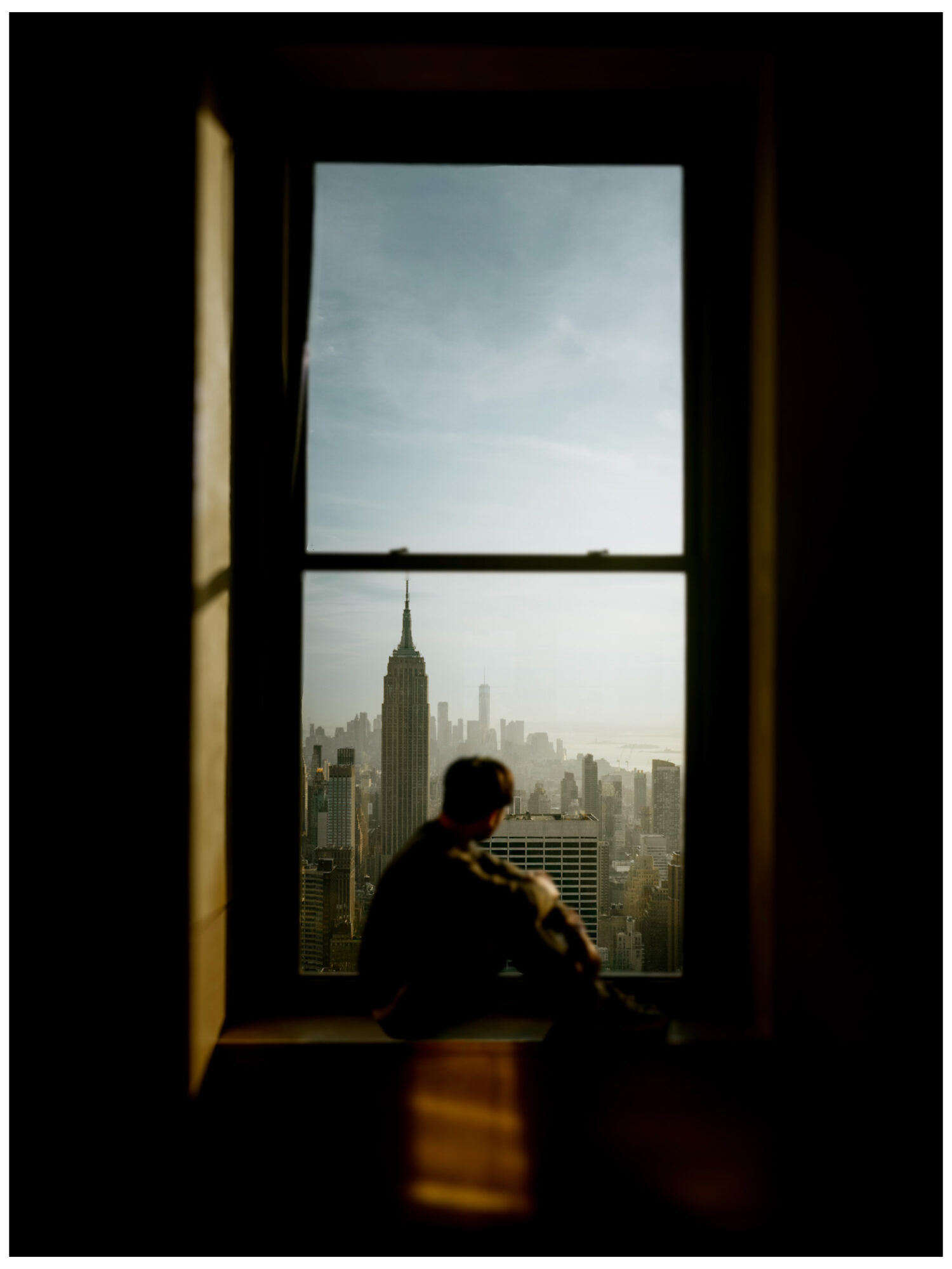Person sitting by a window overlooking a city skyline with tall buildings and a cloudy sky.