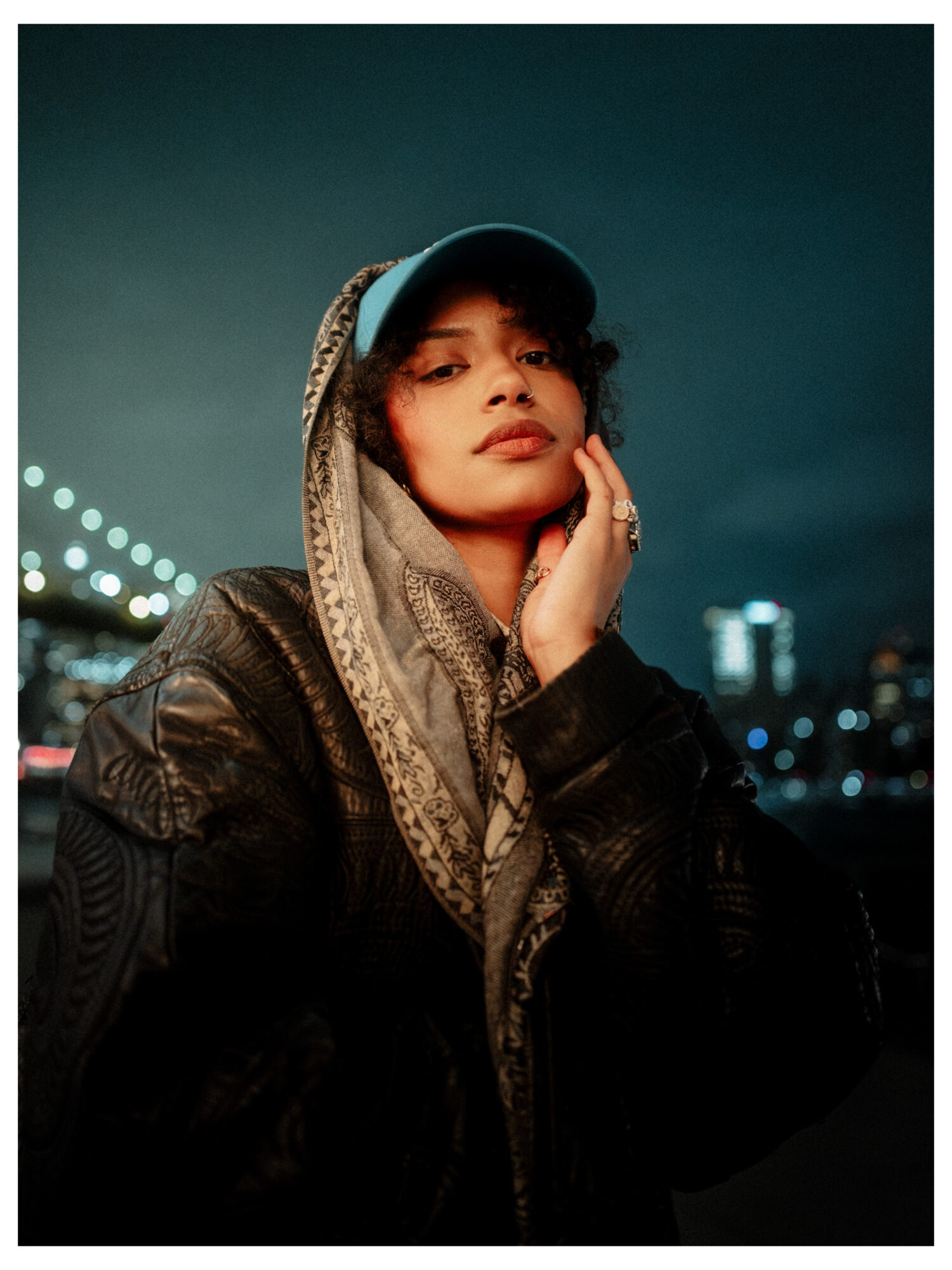 Young woman wearing a cap, scarf, and leather jacket outdoors at night with city lights in background.