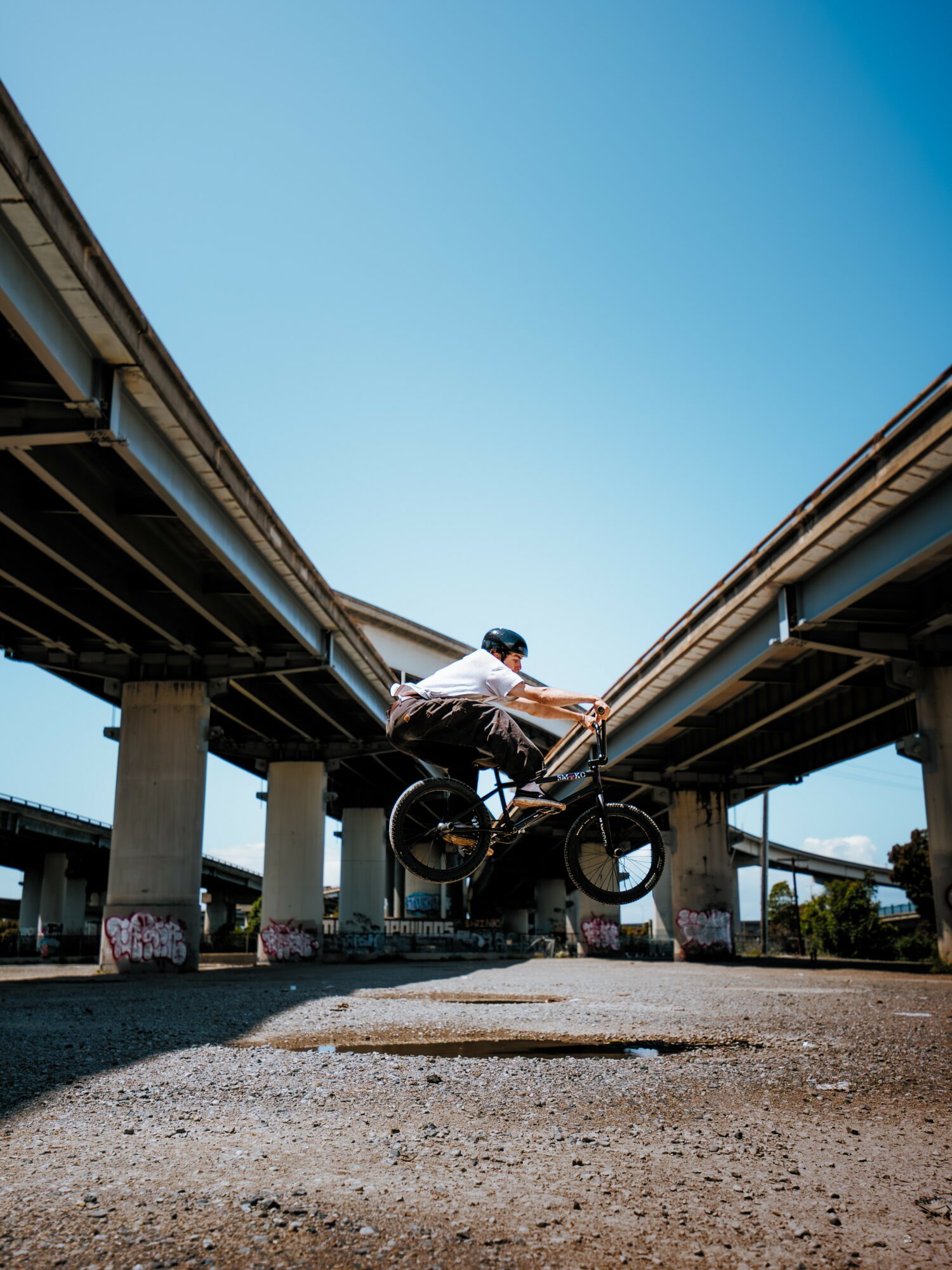 Person riding a bicycle under a bridge, with clear blue sky above, viewed from below.