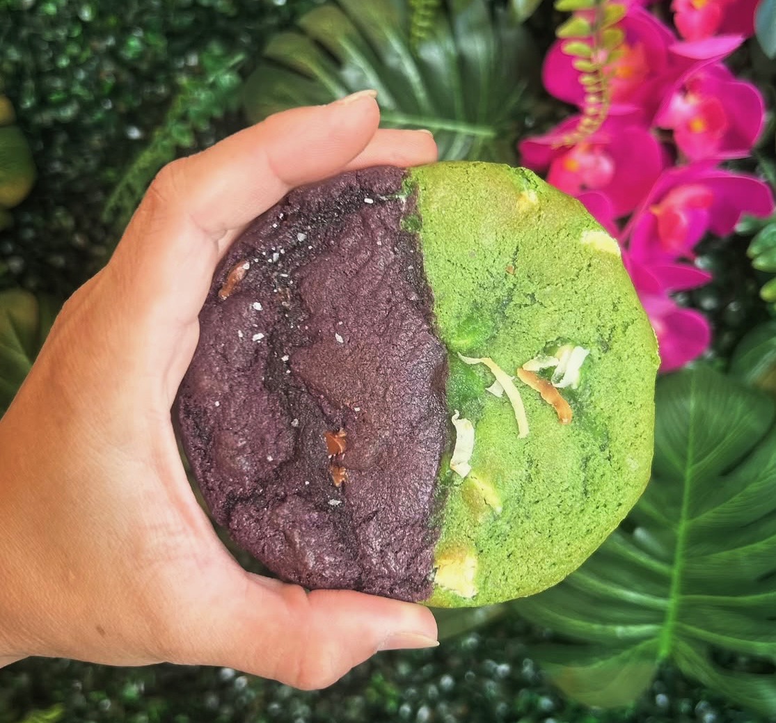 Hand holding a half green and half purple fruit with pink flowers and green leaves in background.