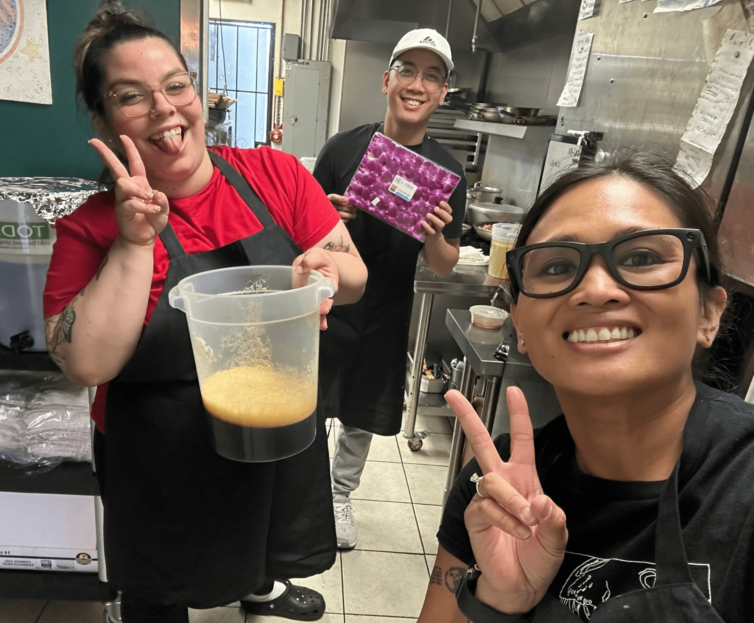 Three people smiling and making peace signs in a kitchen, one holding a container with a yellowish liquid.