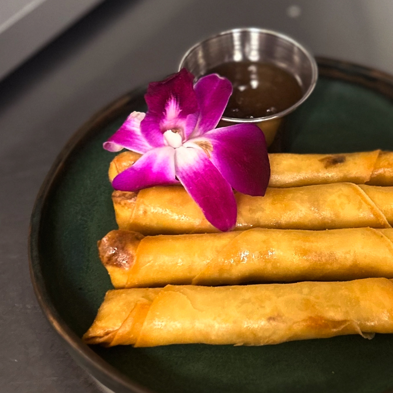 Four golden spring rolls on a dark green plate with a purple orchid flower and a small metal bowl of dipping sauce.
