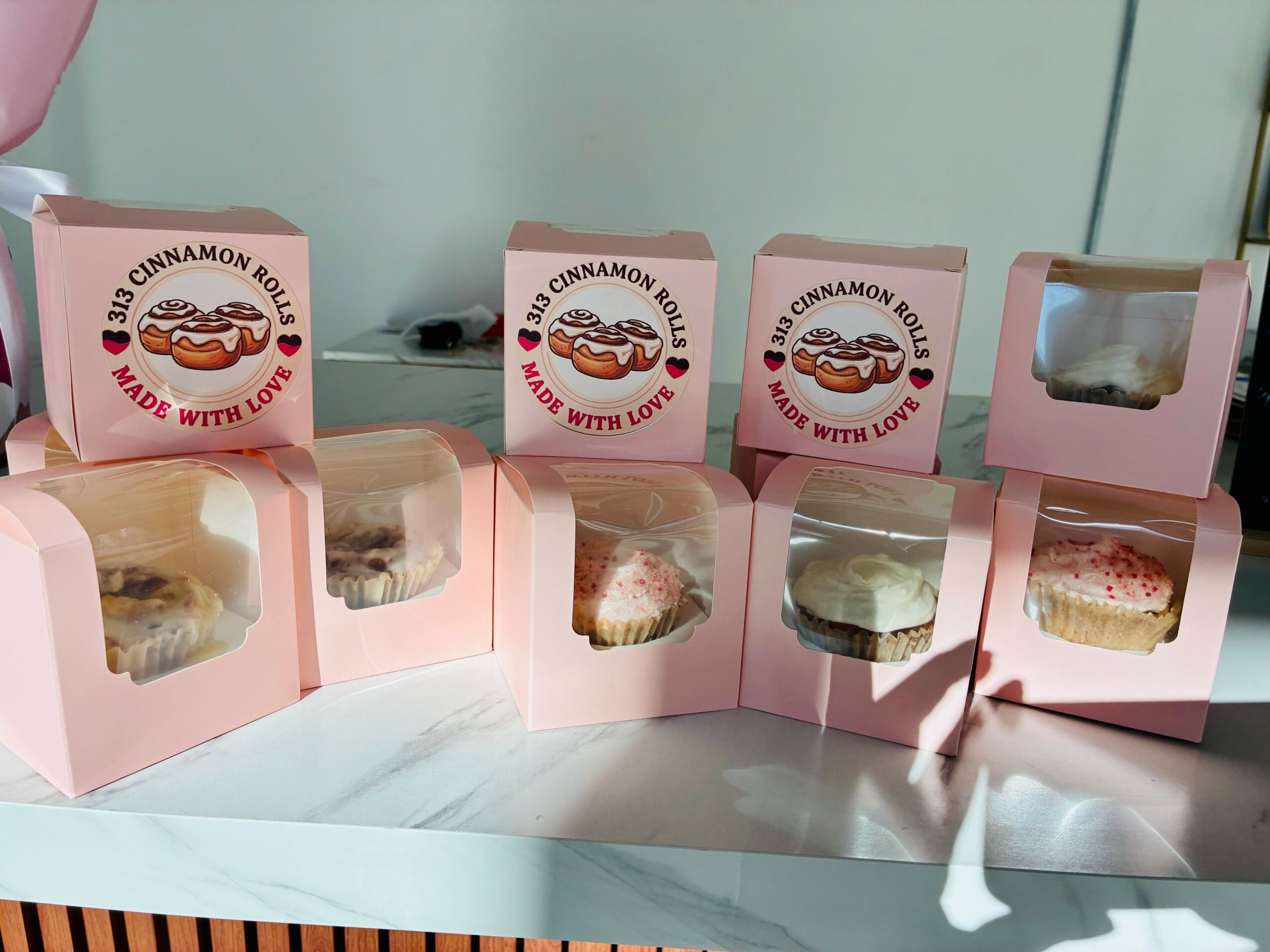 Six pink boxes of cinnamon rolls on a table, some open showing the pastries inside, with a light-colored wall in the background.